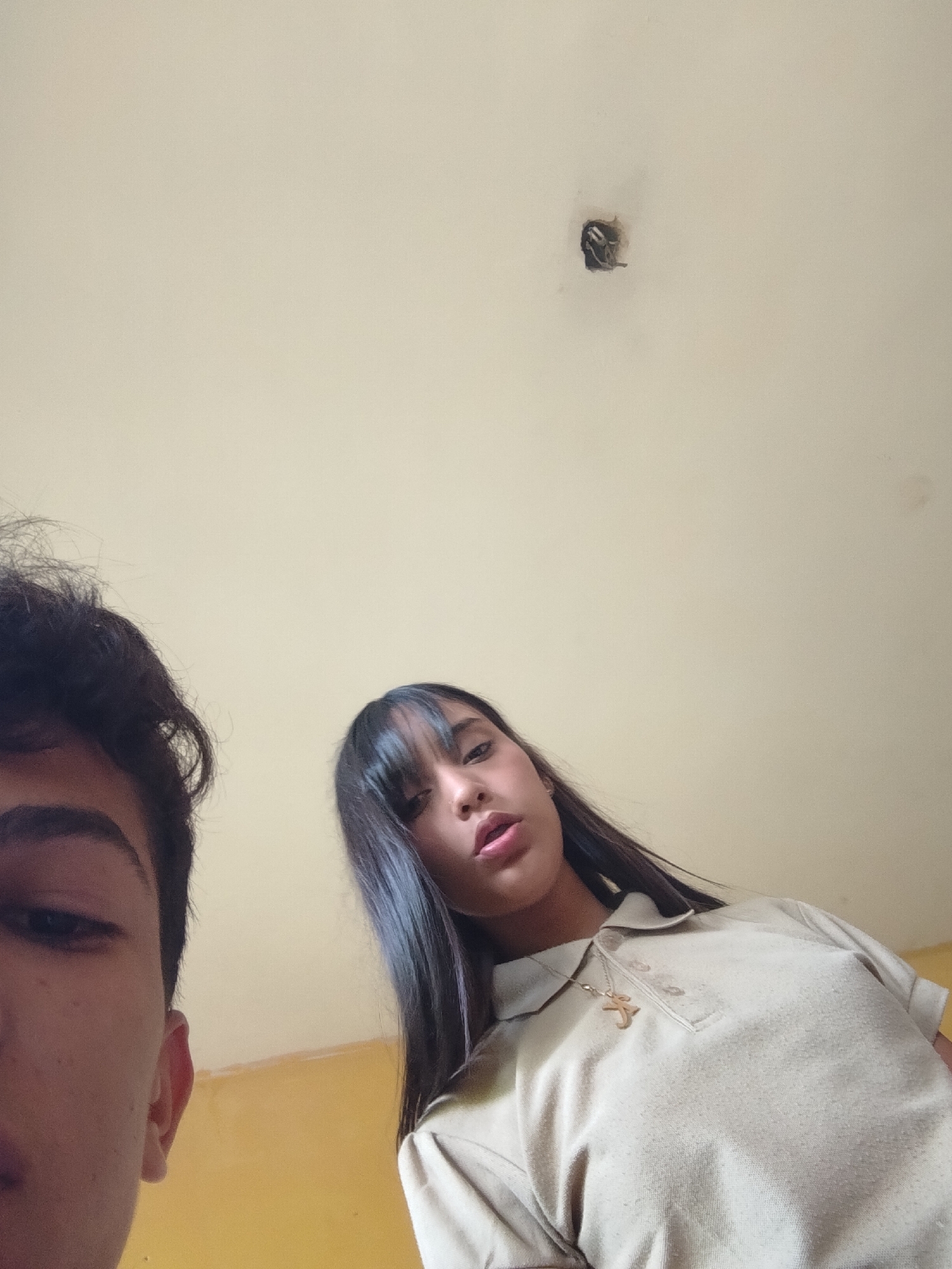 Two young people, a male and a female, are positioned in the foreground of the image, looking up at the camera. The male is on the left, with his face partially visible. The female is on the right, with her face fully in view. She has long, dark hair and is wearing a light-colored polo shirt with a necklace. The background appears to be a ceiling with a light fixture, and a yellow wall is visible at the bottom of the image. The image quality is grainy and taken from a low angle. The specific location within Santa Ana, Venezuela, cannot be determined from the visual information provided in the image.