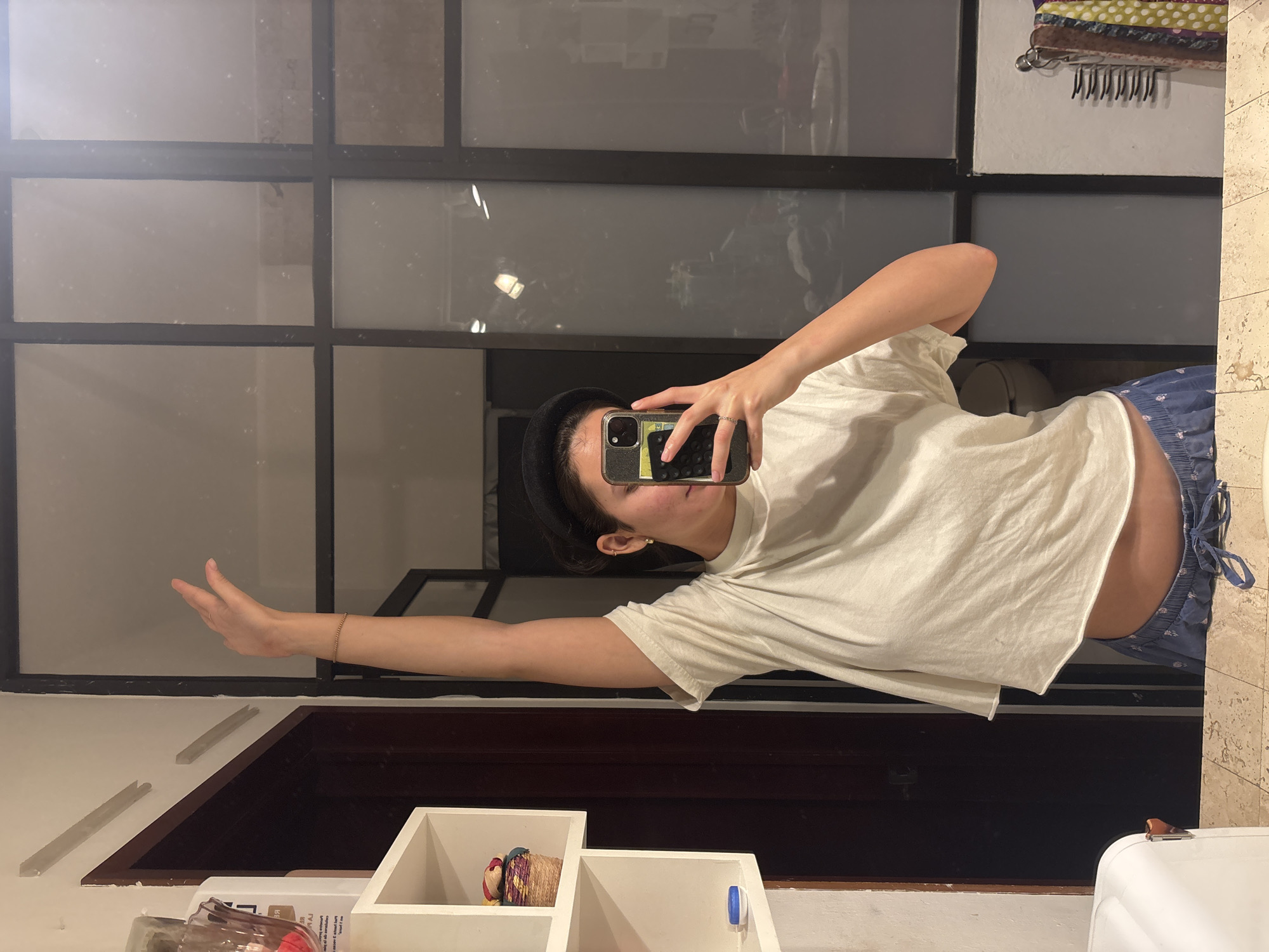 A female is taking a selfie in a bathroom mirror. The subject is wearing a white t-shirt, blue floral-patterned shorts, and a black beret. She holds a smartphone in her right hand, obscuring part of her face. Her left arm is extended towards the left side of the frame. The background consists of a window with a grid pattern, a white wall with a clothes rack, and bathroom fixtures. The setting appears to be a bathroom in Puebla, Mexico.