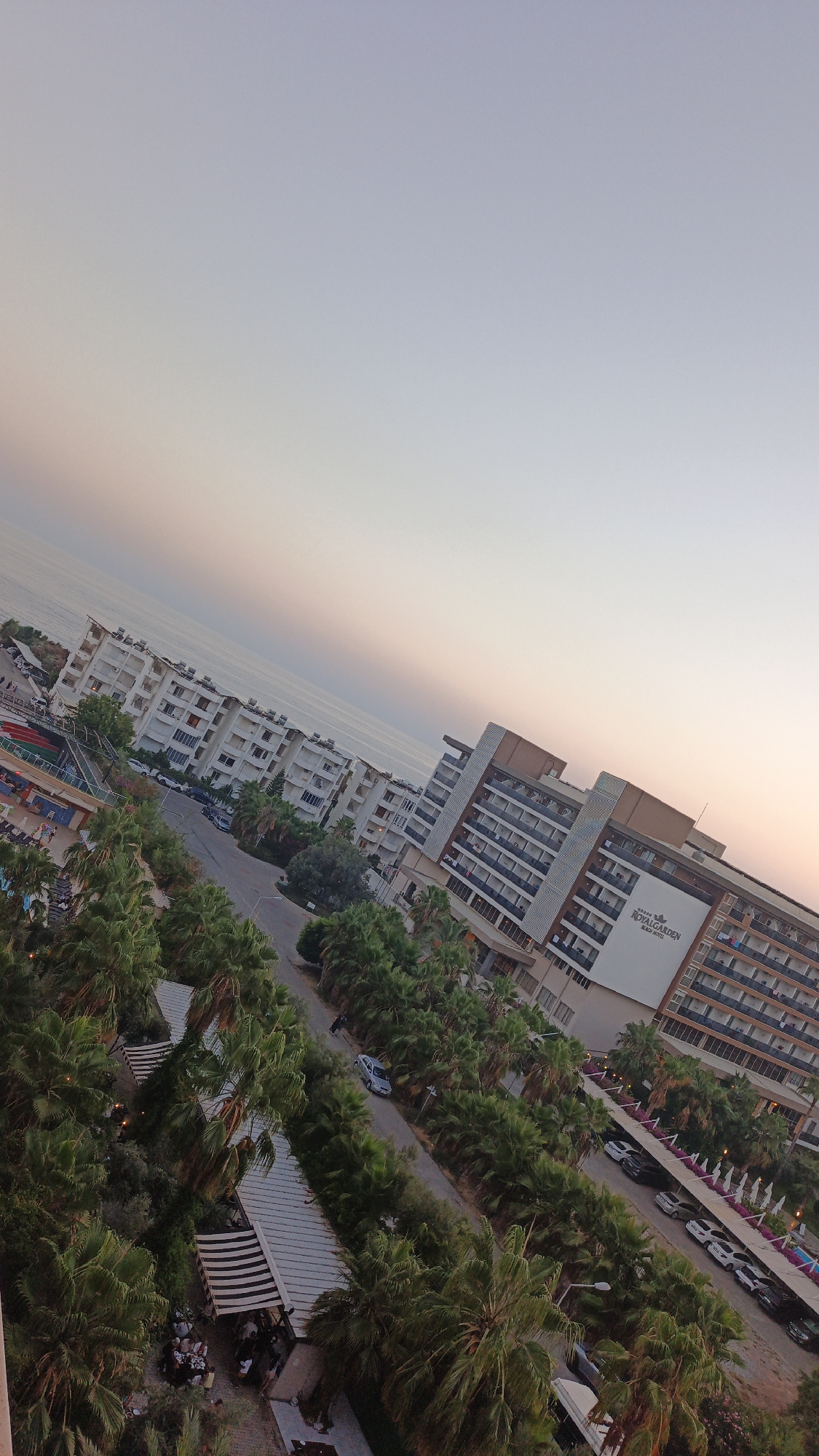 This is an outdoor, high-angle, full shot of a coastal resort area in Alanya, Turkey, taken during the late afternoon or early evening. The sky is a gradient of pale blue to light orange, with the horizon line of the sea visible in the background.
Dominating the mid-ground and right side of the image is a large, multi-story hotel building with a prominent sign reading "ROYAL GARDEN BEACH HOTEL". The hotel has numerous balconies and a modern architectural design. In front of and to the left of this hotel are several other white, multi-story apartment or hotel buildings.
A road runs diagonally through the scene, lined with lush palm trees and other greenery. Several cars are parked along the road and on a lot to the right of the Royal Garden hotel. To the left of the road, there appears to be a swimming pool complex with colorful lounging areas. Further down the road, there are outdoor seating areas with striped awnings, where small groups of people are gathered at tables.
A single person is visible walking along the road in the mid-ground. The overall impression is one of a bustling resort environment, with a focus on leisure and accommodation. The image is slightly tilted.