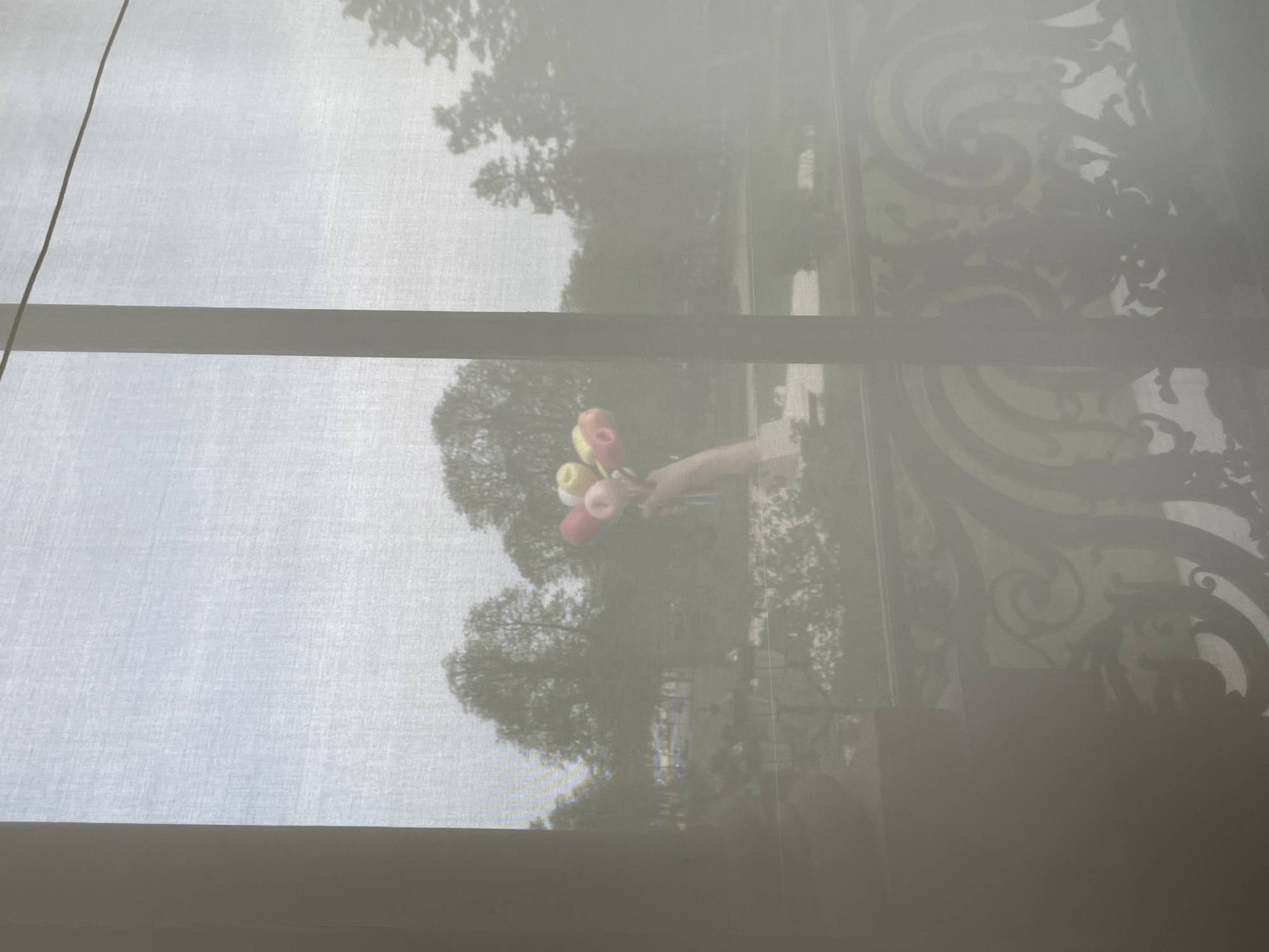 The image presents an outdoor scene viewed through a translucent, white fabric window covering. A light-skinned human hand, positioned in the lower-mid-left, holds a cluster of five pastel-colored spherical objects (pink, red, yellow, and light). Immediately beyond the window, an ornate, dark metal balcony railing with intricate scrollwork spans the lower portion of the frame. The external environment depicts a public park or garden in Paris, France, characterized by numerous mature trees under an overcast sky. A multi-lamp street light is faintly visible in the mid-ground, and in the distant left, a bright white structure with yellow accents, possibly a vendor stand, is discernible through the trees. A dark vertical window frame element bisects the view.