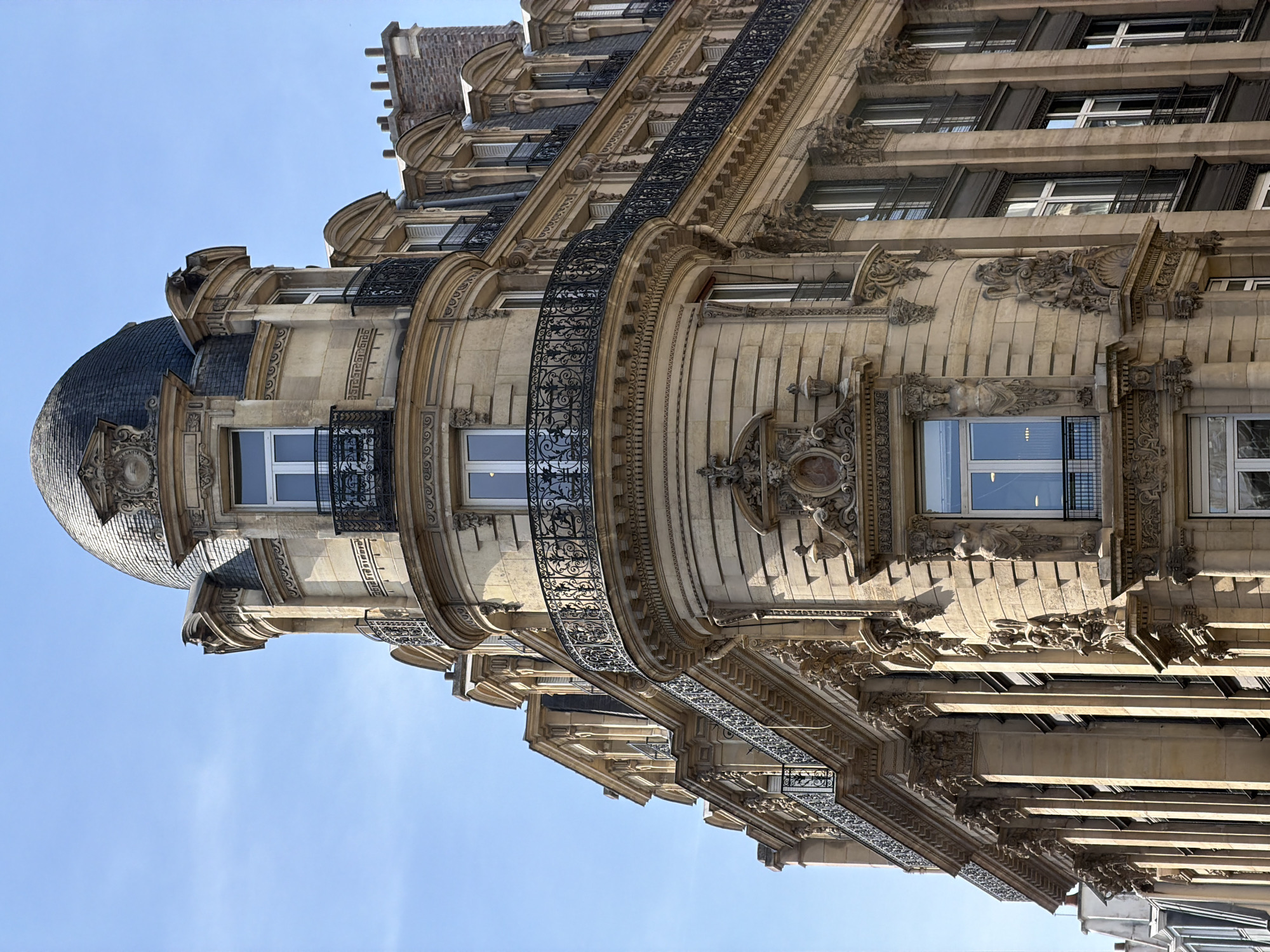 The image shows a detailed view of a Parisian building facade. The building features elaborate architectural details, including ornate stone carvings, wrought-iron balconies, and a domed corner turret. The building's style is classical, with elements of the Beaux-Arts architectural style. The facade is made of light-colored stone, possibly limestone, which is typical of Parisian architecture. The windows have dark shutters. The sky is clear and blue. The image is taken from a low angle. The building is located in Paris, France, recognizable by the architectural style.