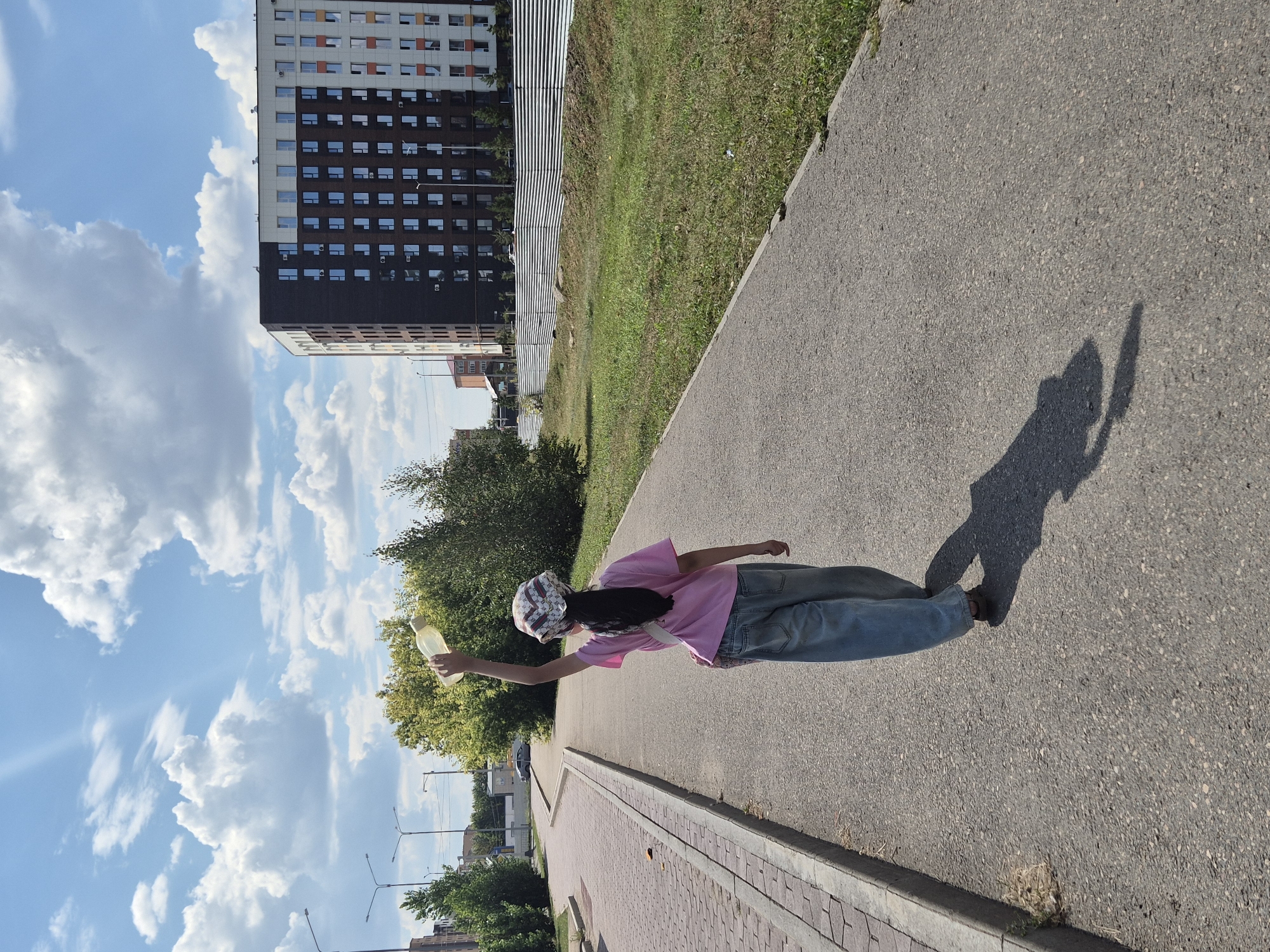 A young person, viewed from behind, walks on a paved path in Kokshetau, Kazakhstan. They are wearing a pink t-shirt, wide-legged blue jeans, and a patterned headscarf. The person holds a light-colored plastic water bottle above their head with their right hand. A dark shadow is cast on the path by the person and the bottle. To the left of the path is a sidewalk made of brick pavers, separated by a concrete curb. Beyond the sidewalk are trees and a multi-story residential building with a dark brown and white facade, featuring numerous windows. To the right of the path is a grassy area bordered by a corrugated metal fence. The sky is blue with scattered white clouds. The overall scene depicts a bright, sunny day.