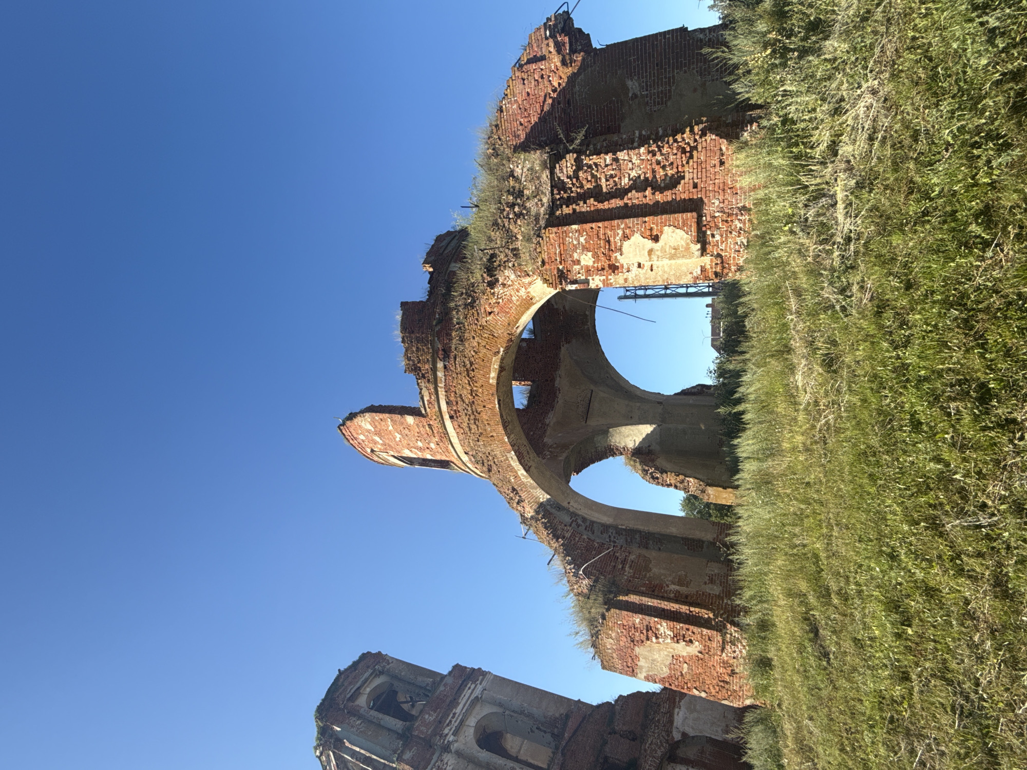 The original image shows the ruins of a brick structure against a clear blue sky. The structure is partially collapsed, with arched openings and visible brickwork. Some vegetation, likely grass and weeds, is growing on and around the ruins, and also covers the foreground. To the left of the main structure is a tall, slender brick tower, also in a state of disrepair. The overall impression is one of abandonment and decay. The scene is outdoors, with no people or animals visible. The lighting suggests it is daytime, likely morning or afternoon due to the bright, clear sky. The location is described as Unknown, Unknown.