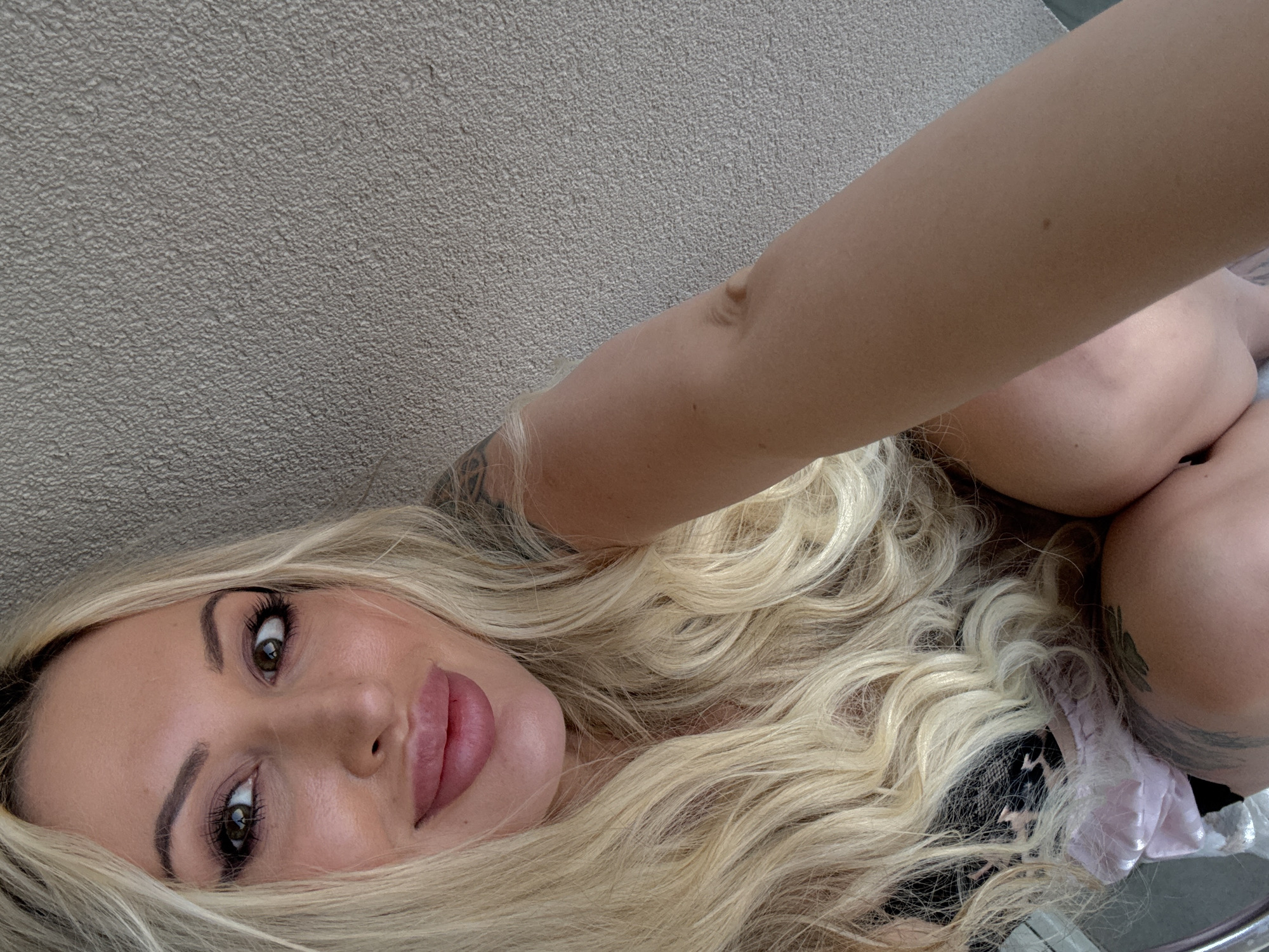 A woman with long, wavy blonde hair and makeup is shown in a close-up shot. She is wearing a black garment and a pink, silky fabric is visible. Tattoos are present on her arm and leg. The background appears to be a textured, light-colored wall. The setting is not discernible.