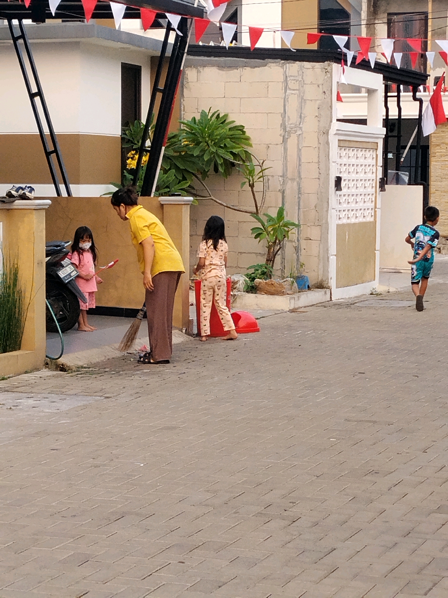 In the city of Sukabakti, Indonesia, a woman wearing a yellow shirt and brown pants is sweeping the paved ground with a broom. She is facing away from the camera. To her left, a young girl in a pink outfit and mask stands near a motorcycle. To the woman's right, another young girl in a patterned pink outfit stands. Further to the right, a young boy in a blue and black patterned shirt and shorts walks away from the camera. Red and white triangular flags are strung across the top of the frame, indicating a celebration or event. The scene takes place on a street with houses in the background.