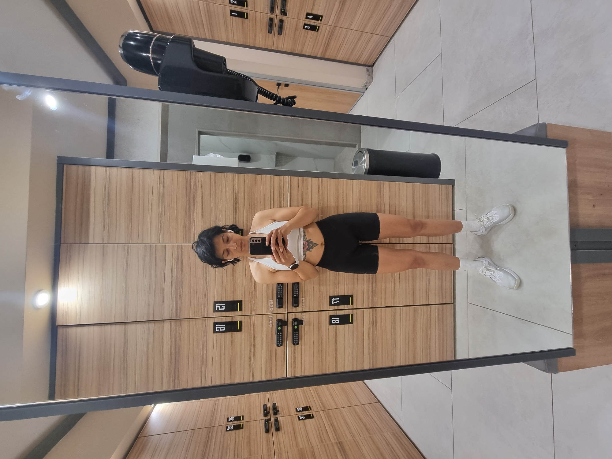 A woman is reflected in a mirror in what appears to be a gym locker room. She is wearing a white tank top and black biker shorts, holding a black smartphone to take a selfie. She has short dark hair and is wearing white sneakers. The background consists of a wall of wooden lockers with black hardware. There is a hairdryer mounted on the wall above the lockers, and a black trash can next to the wall. The floor is tiled in light grey. The location in Plovdiv, Bulgaria, cannot be determined from the image.