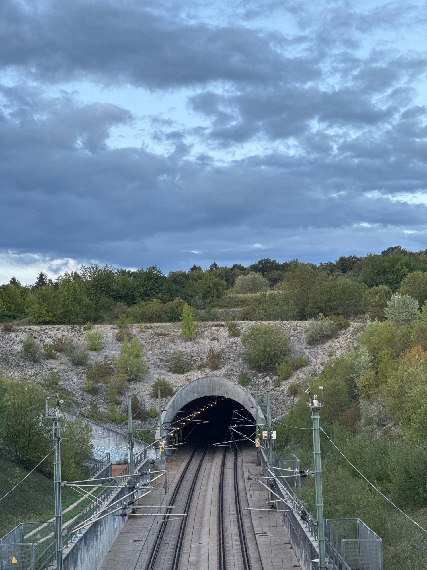 The image shows a view of a train tunnel entrance from an elevated perspective. Twin railway tracks lead directly into the tunnel, which is illuminated by several lights. The tunnel is carved into a hillside covered with gravel and sparse vegetation, with a dense forest visible at the top of the hill. The sky above is overcast with dramatic, dark clouds. Electrical lines and support poles for the railway are prominent in the foreground and along the tracks. The scene appears to be located on the outskirts of Wettstetten, Germany, specifically near the ICE tunnel. There are no people or animals visible in the image.