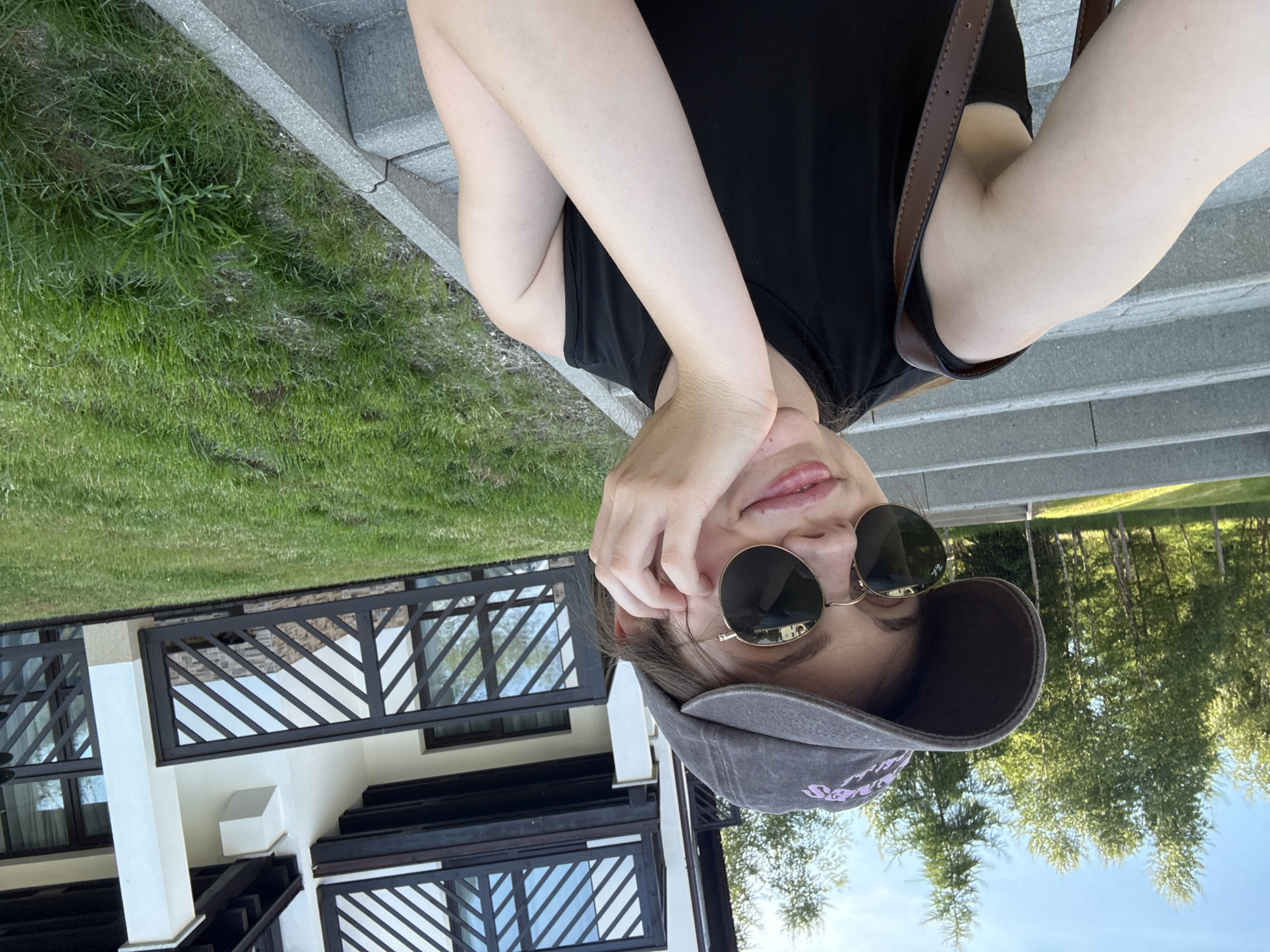 The image shows a young woman with fair skin, wearing a dark baseball cap and sunglasses. She has her right hand resting on her cheek and is looking at the camera with a slight smile. She is wearing a black sleeveless top and a brown strap from a bag is visible over her left shoulder. The woman is sitting on outdoor stairs made of concrete. Behind her, there is a green grassy area and a building with a modern facade featuring dark wooden railings and balconies. Trees are visible in the background, suggesting an outdoor setting, possibly a resort or hotel. The sky is blue with some clouds. This scene is located on the grounds of an apartment building in Szklarska Poręba, Poland.