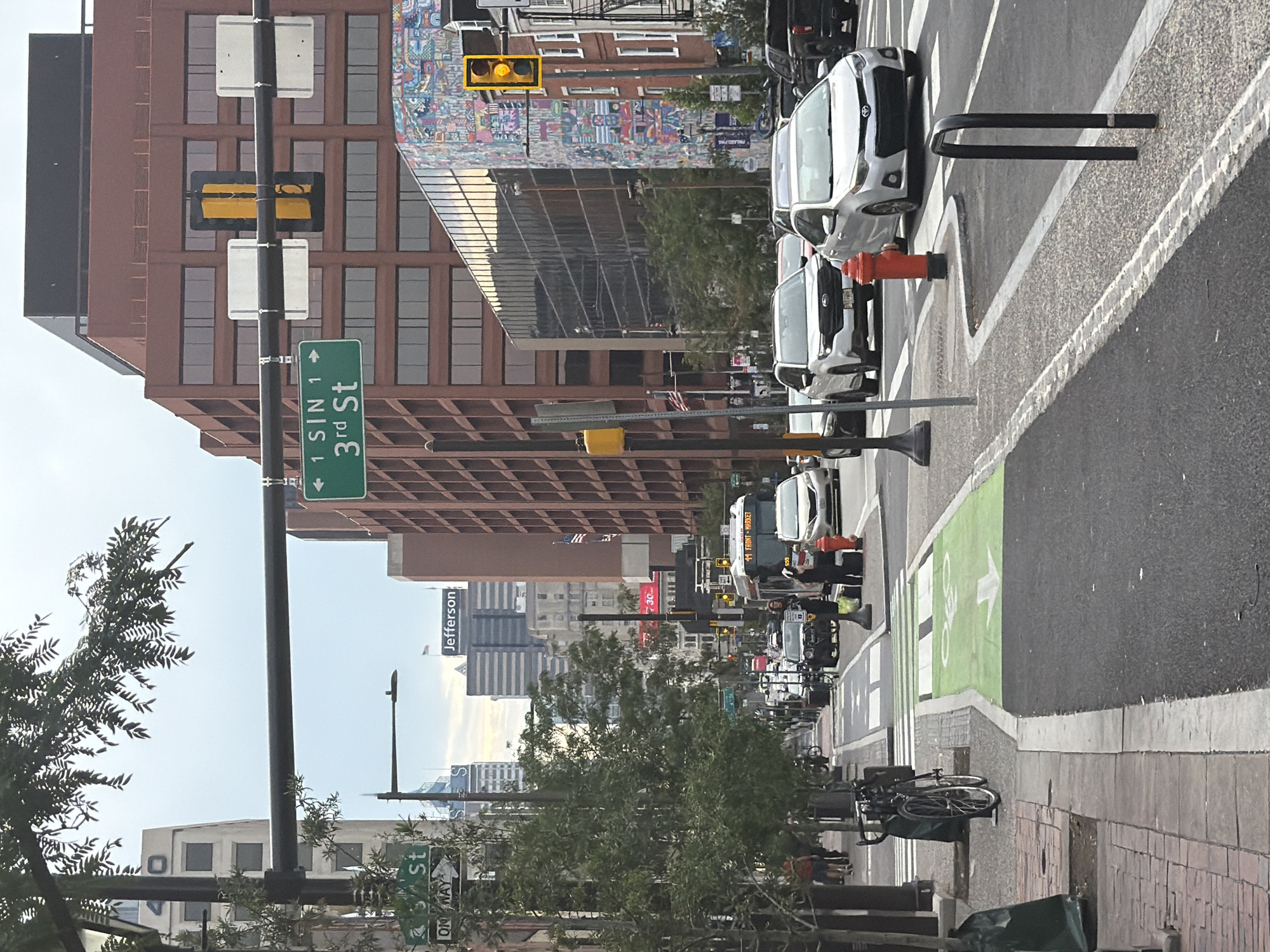 The image captures a street scene in Philadelphia, Pennsylvania, United States of America. The perspective is from a street-level vantage point looking down a city street lined with buildings and trees. In the foreground, a bike lane is visible, marked with green pavement and a bicycle symbol. A parked bicycle rests on the sidewalk to the left. Several vehicles, including cars and a bus, are proceeding down the street. Pedestrians are present on the sidewalks. Traffic lights and street signs are visible, indicating traffic management. Buildings of varying architectural styles, some with reflective glass facades and others with brickwork, define the urban environment. A mural adorns the side of one building, adding a splash of color to the cityscape. The sky is overcast, suggesting it might be a cloudy day. The overall scene depicts a typical urban street environment in Philadelphia.