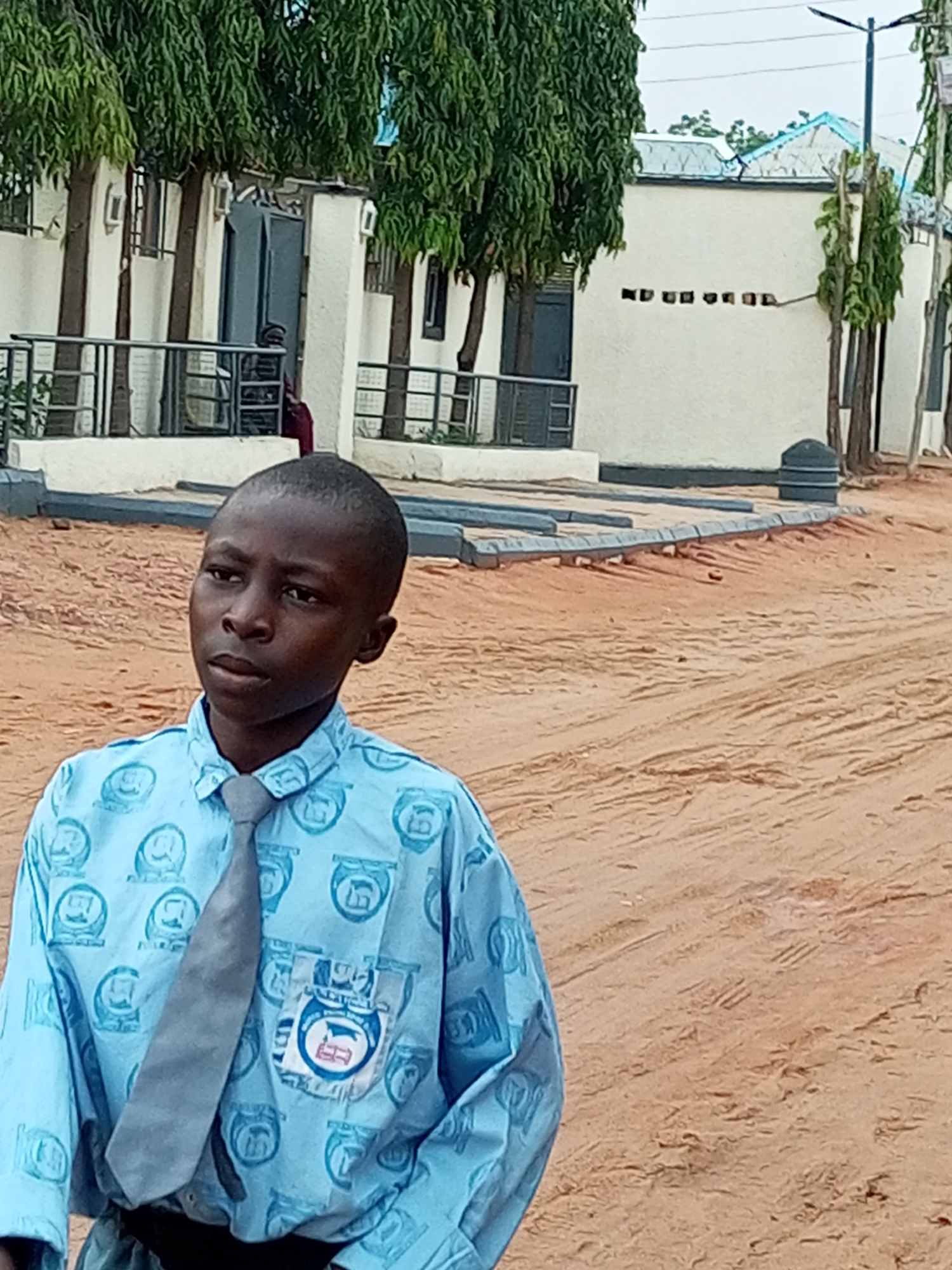 A young boy stands in front of a building with trees in the background. The boy is wearing a light blue shirt with a repeating circular emblem and a grey tie. He has short dark hair and is looking to his left. The building behind him is white with dark doors and windows, and a metal railing runs in front of it. The ground is made of light brown dirt, with tire tracks visible. The sky is overcast.