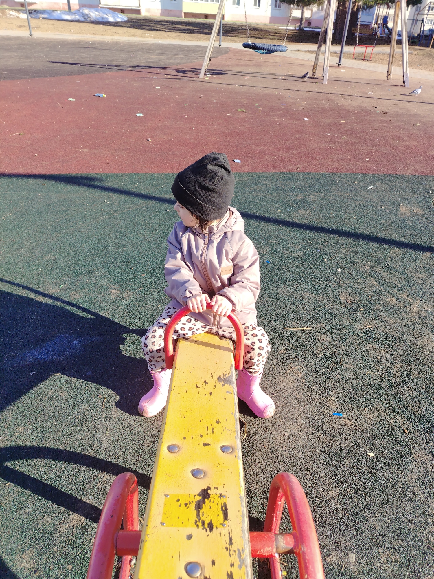 A child, approximately 3-5 years old, sits on a yellow and red seesaw at a playground. The child is wearing a black beanie, a light brown jacket, leopard print pants, and pink boots. The playground surface is green and red. In the background are swing sets, and a snow-covered area can be seen. This appears to be in Noviy, Russia.