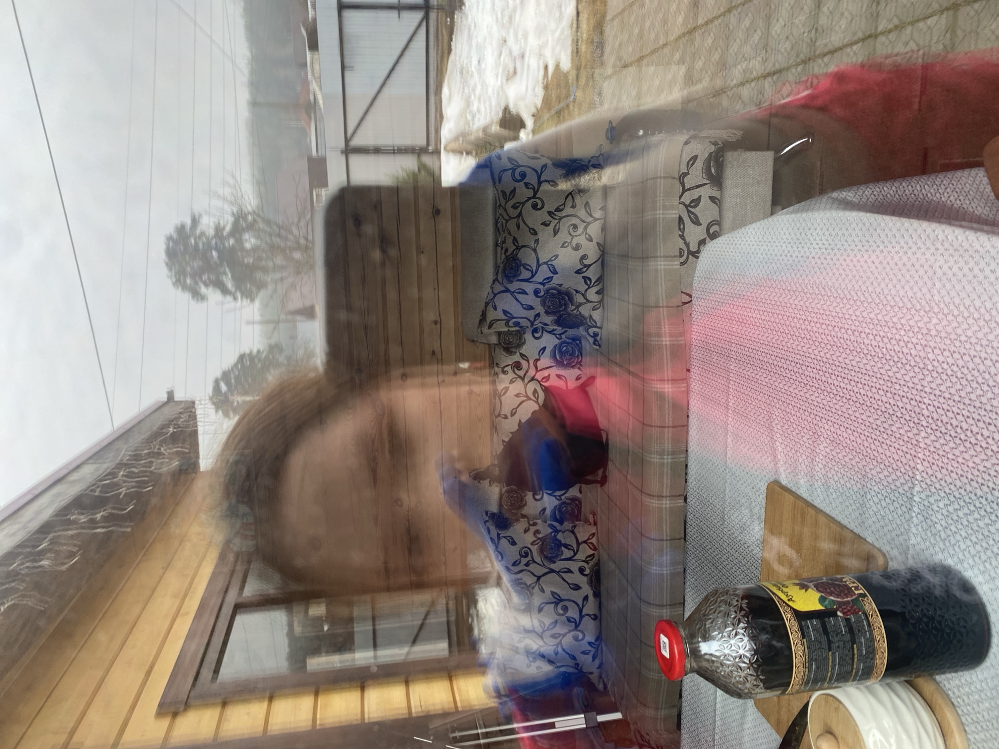 The image appears to be a reflection in a window. The reflection shows a person with brown hair, a building with wooden siding, a snowy outdoor scene, and interior elements. Inside, there's a couch with floral and plaid patterned pillows, a red garment, and a table with a white textured surface. On the table, there's a bottle with a red cap, a wooden stand, and a white spool. Without further data it is not possible to verify if this picture has been taken in Chaikovsky, Russia.