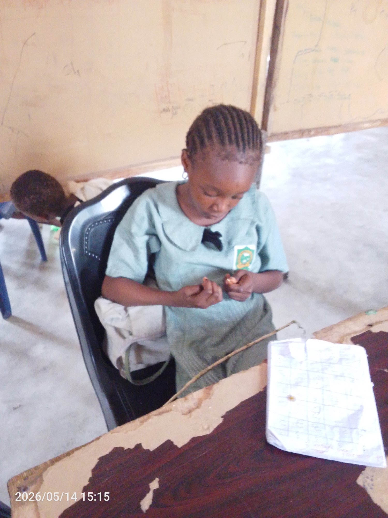 A young girl is seated at a desk in what appears to be a classroom, possibly in Lokoja, Nigeria. She is wearing a light green uniform with a school emblem. Her hair is braided. She looks down at something in her hands. On the desk, there is a piece of paper with lines drawn on it. The room has bare walls. A timestamp of "2026/05/14 15:15" is displayed. Another child is sitting in the background.