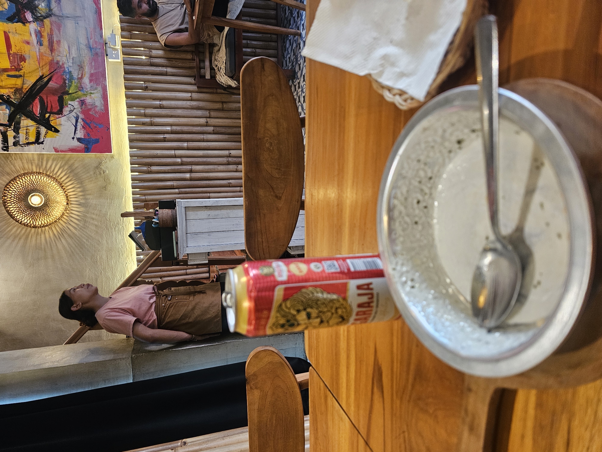 In a cafe in Ubud District, Indonesia, a young woman in a pink t-shirt and brown apron stands leaning against a pillar, looking to her right. In the foreground, a can of "Brahma" beer sits on a wooden table next to a bowl of food with a spoon. In the background, bamboo walls line the space, and a man with a beard is seated at a table. Abstract art hangs on the wall above.