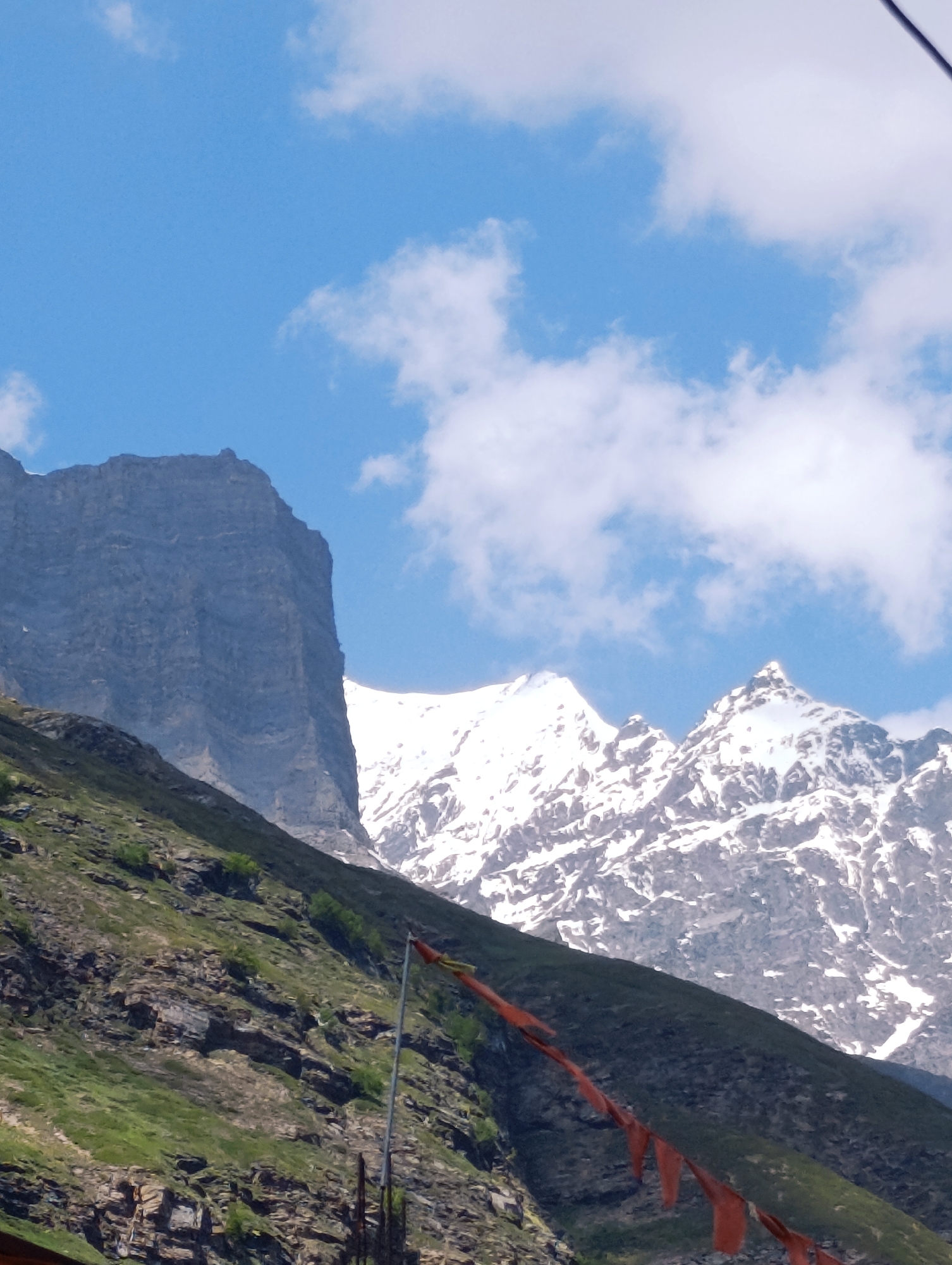 This is a vertical, eye-level shot of a mountainous landscape in Yamling, India. The scene is dominated by towering, rugged mountains, some of which are capped with snow. The sky is a bright blue with scattered white clouds.
In the foreground, a steep, rocky slope is covered in patches of green grass and vegetation. A string of red prayer flags is visible, draped across the hillside and extending towards the right of the frame. The flags are slightly faded and appear to be made of cloth. A tall, thin pole, possibly a flag or prayer flag pole, is also present in the lower right quadrant of the image.
The mountains themselves are imposing. A massive, dark gray rock face with distinct horizontal striations rises on the left side of the frame. To the right of this, snow-covered peaks jut sharply into the sky. The lighting suggests it is daytime, likely late morning or early afternoon, with the sun casting shadows on the mountains. The weather appears clear and pleasant.
There is no visible text in the image, nor are there any people or specific activities depicted. The overall impression is one of natural grandeur and a sense of peaceful, remote wilderness. The prayer flags hint at a spiritual or cultural significance of the location.