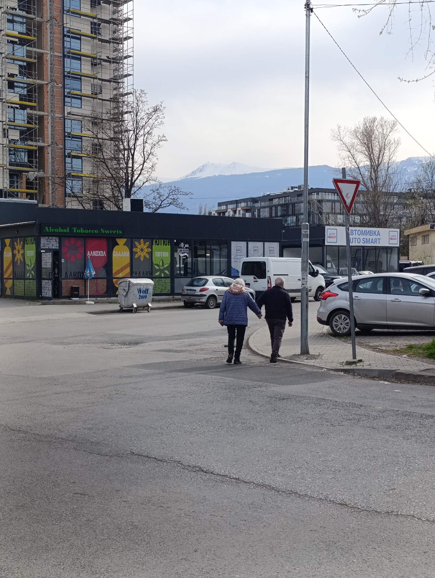 The image shows a street scene in Sofia, Bulgaria. In the foreground, a man and a woman are walking hand-in-hand across the street. The man is wearing a dark jacket and gray pants, while the woman is wearing a blue jacket and black pants. They are walking away from the camera. To the left of the couple is a building labeled "Alcohol Tobacco Sweets". On the right of the image is a building labeled "AUTOMIVKA UTO SMART". Several parked cars are visible in the background. In the background, there are other buildings. A tall building under construction is also visible in the background on the left side of the image. In the distance, there are snow-capped mountains. A utility pole with street signs is on the right side of the image.