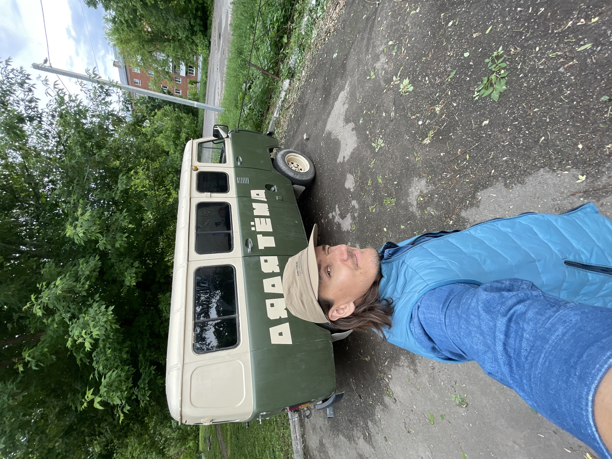 A man wearing a blue vest and a beige cap stands next to a vehicle with the inscription "АЯЯ ТЕМА" on its side. The vehicle is green and beige. The location is outdoors on a paved surface with patches of vegetation and fallen leaves. In the background, there are trees, a utility pole, and a multi-story building. The orientation of the image and inscription suggest the photo was taken with the subject lying on their side. While the city is not visible in the image, it can be inferred from the inscription on the vehicle that the location is likely in Russia.