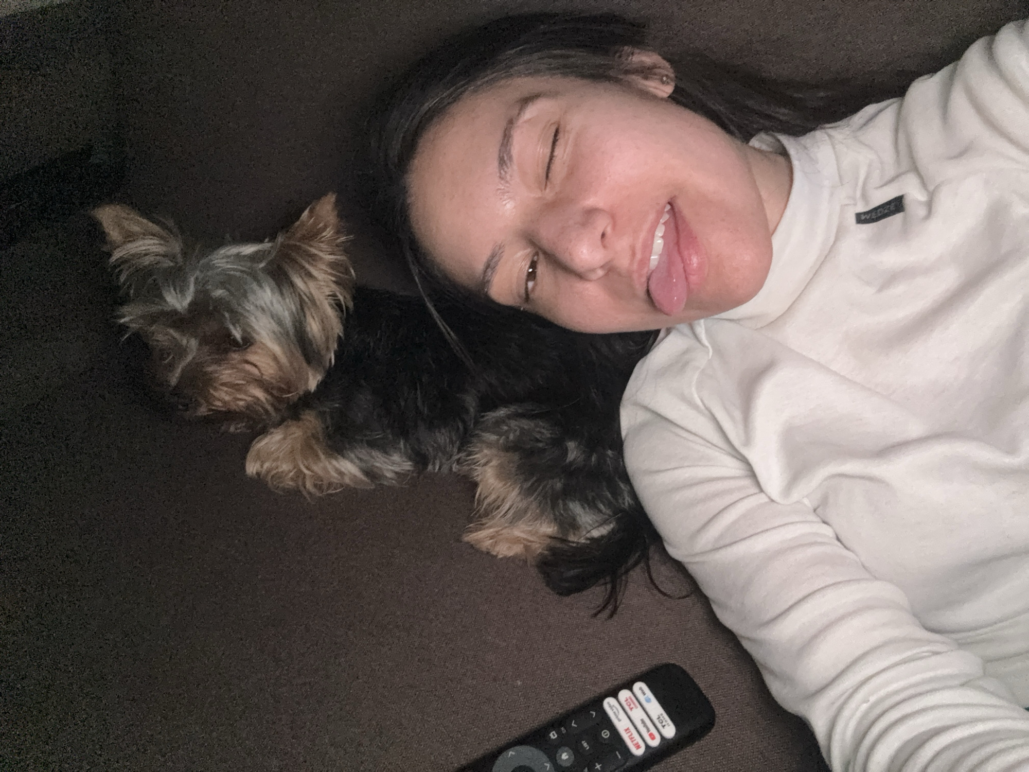 A woman is lying on a dark brown couch with a small dog. The woman is fair-skinned and has dark hair, and she is sticking her tongue out with one eye closed. She is wearing a long-sleeved white shirt. Next to her, to the left, is a small Yorkie dog lying down. Below the dog is a black TCL Roku TV remote. The location is not determinable to be Varna, Bulgaria.