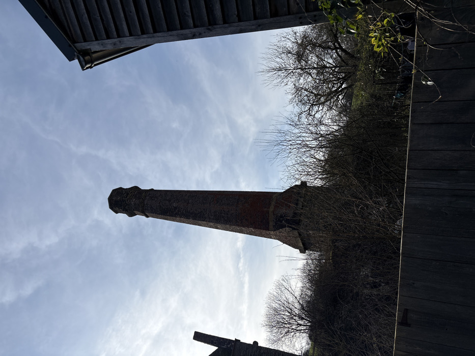 The image presents an outdoor scene at Stiperstones, United Kingdom, under a partly cloudy sky. A prominent, tall industrial chimney constructed of reddish-brown brick stands centrally, tapering from a broad, multi-faceted base to a narrower shaft, culminating in a stepped, crenellated cap. To the far left, a partial, dark brick structure is visible. On the far right, a dark, wooden-clad building with horizontal siding and a visible dark gutter beneath its roofline is partially framed. The lower foreground is occupied by a dark wooden fence composed of horizontal planks. Between the fence and the structures, dense, tangled bare deciduous trees and brush are heavily shadowed. Beyond the trees, a distant grassy area is visible. Faint, light-colored shapes consistent with laundry drying on a line are discernible within the bare trees on the right. No individuals are present.