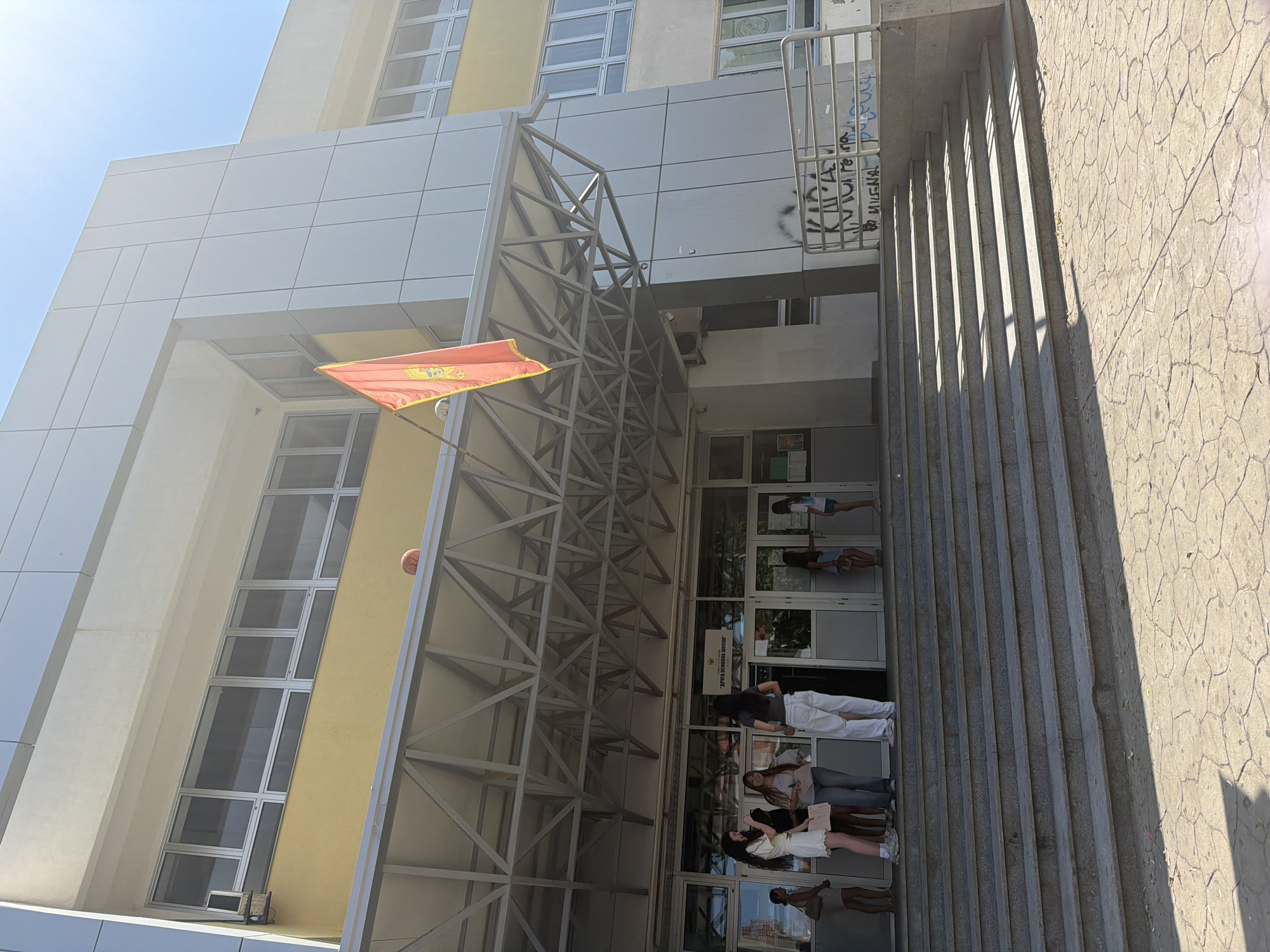 The image depicts the exterior of a modern building, likely a school, with a light yellow and white facade. A prominent feature is the metal truss structure extending over an entrance. A flag with a red background and a golden crest is visible, suggesting the location might be Montenegro, as indicated by the provided context. Sunlight casts long shadows on the textured stone steps leading up to the entrance, indicating it is daytime, likely late afternoon.
Several people are seen near the entrance. Some are standing on the steps, appearing to be students, some holding books or papers. Their postures and expressions are neutral. A basketball is visible resting against the side of the building. The windows of the building are large and reflective, showing glimpses of the interior. Some graffiti is present on the wall near a metal railing. The overall scene is bright and airy, suggesting fair weather. The building's architecture is modern, with clean lines and large glass elements. A sign with Cyrillic text is affixed to the entrance doors.