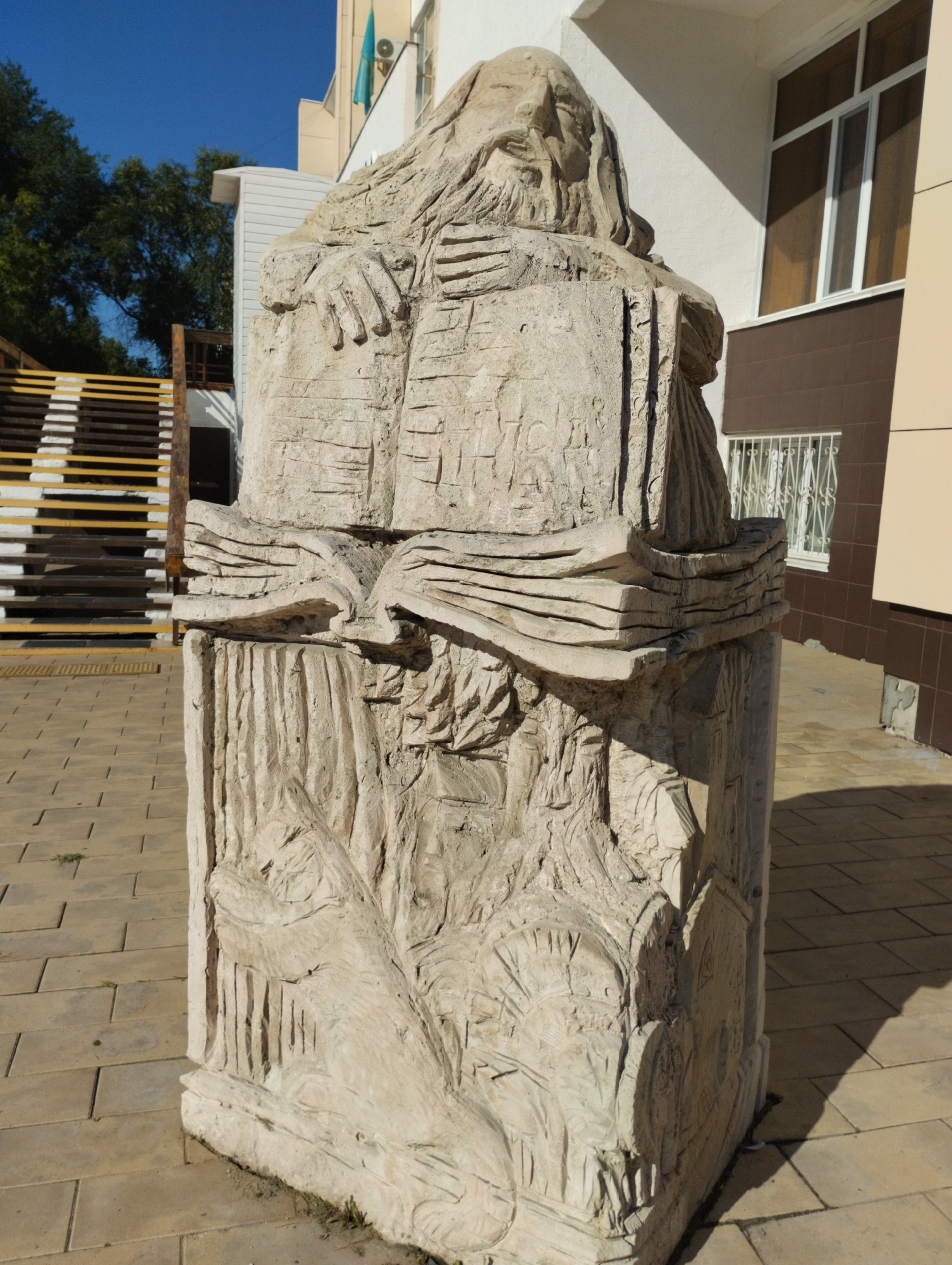 A light-colored stone sculpture of a seated, robed figure reading a book stands on a paved sidewalk. The sculpture has a long beard and hair, and its hands are resting on the open book. The lower part of the sculpture is carved with vertical lines. In the background, a modern building with brown and white siding and a window is visible. To the left of the sculpture, a set of outdoor stairs with yellow and white stripes leads up. A blue flag with a gold emblem, likely the flag of Kazakhstan, is attached to the building. The sky is clear and blue. This sculpture is located in Kostanay, Kazakhstan, in front of the Kostanay State Pedagogical Institute.