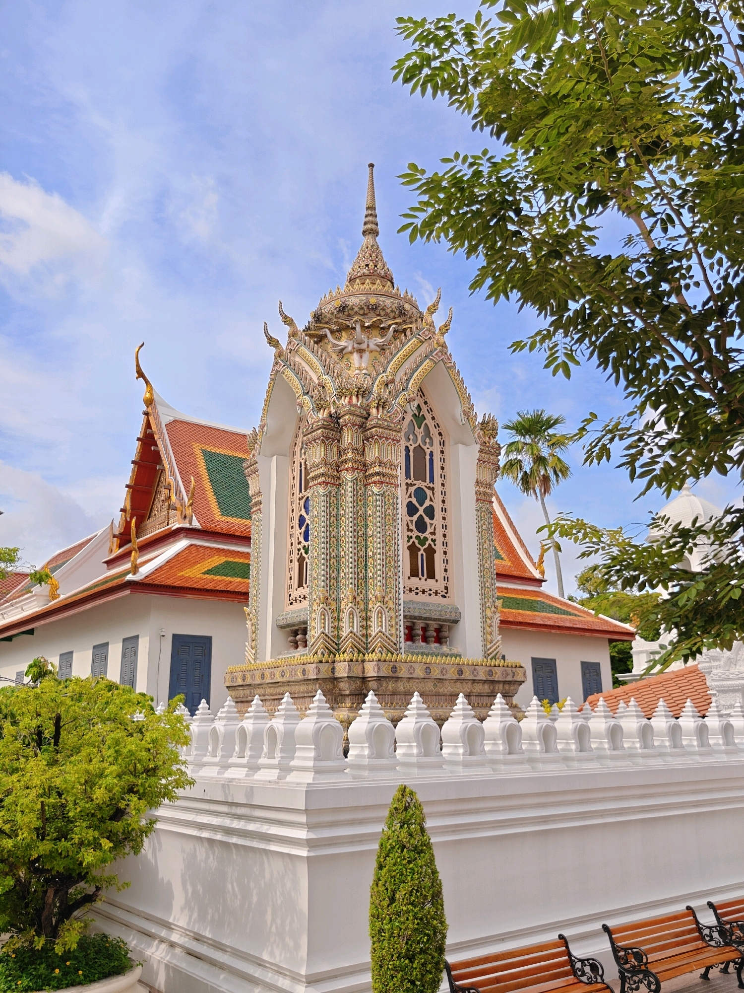 This image depicts a vibrant, ornate Buddhist temple structure, likely a pavilion or shrine, situated within a temple complex in Bangkok, Thailand. The structure is a multi-tiered, pagoda-like building adorned with intricate mosaic tiles in shades of gold, green, blue, and white. It features decorative spires and arched windows with stained glass detailing. To the left, a larger temple building with a traditional Thai roof, featuring orange and green tiles and gilded trim, is visible. A white, low wall with decorative finials runs along the foreground, separating the temple grounds from an area with wooden benches. Lush green trees and foliage frame the scene, with a tall palm tree visible in the background. The sky is partly cloudy and blue. There are no people visible in the image. The scene conveys a sense of religious and architectural grandeur characteristic of Bangkok's temple landscape.