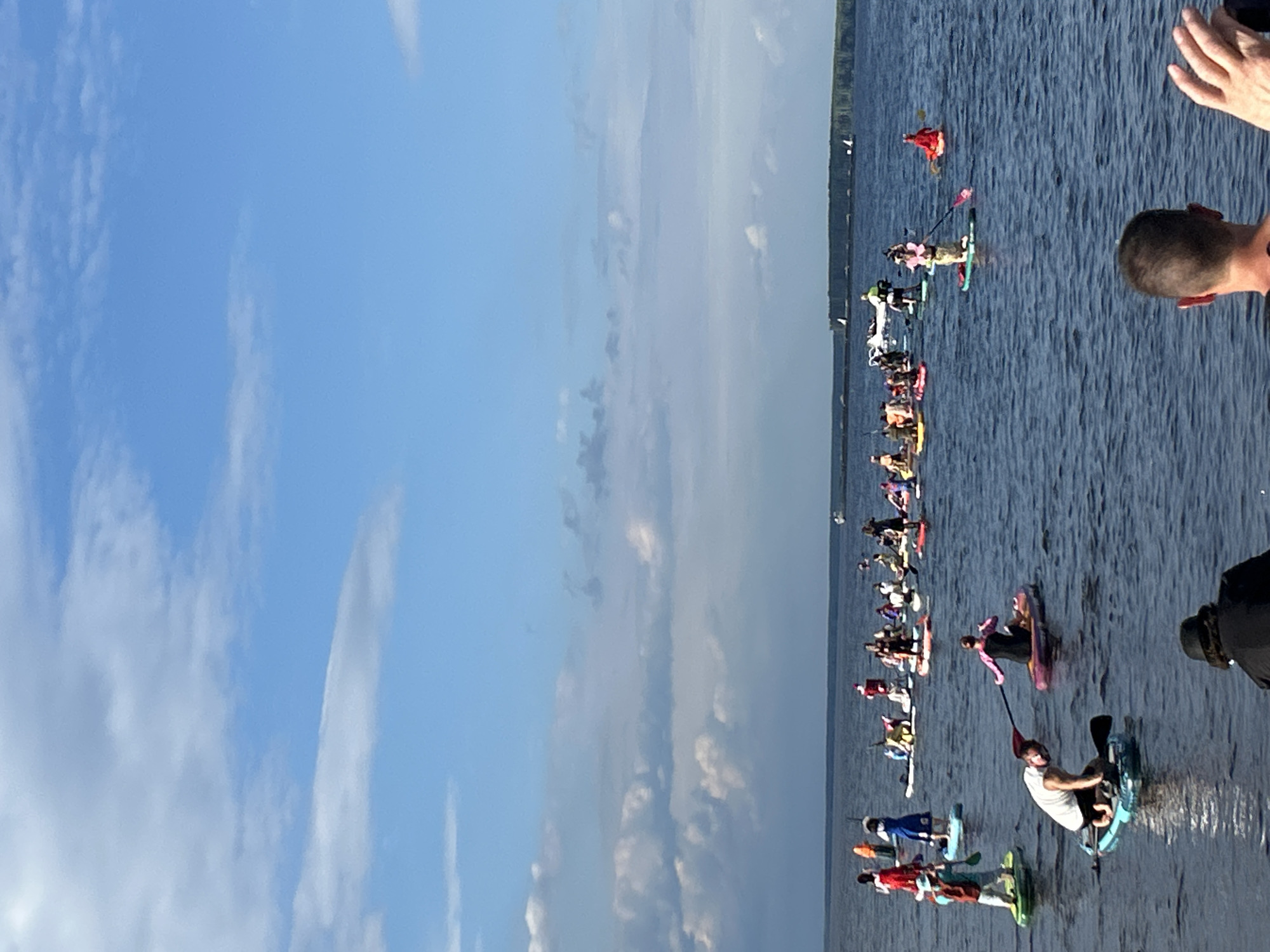The image depicts a sunny day with a clear blue sky dotted with scattered clouds over a body of water. A group of people are paddleboarding on the water, some in costume. There are approximately 30 paddleboarders in the frame, spread out across the water. The paddleboarders are varied in age and gender, and some are wearing hats and colorful clothing, possibly for a festive event. Some are paddling actively, while others appear to be posing or resting. In the foreground, two individuals are visible from behind, with one holding a phone, possibly taking a picture. A distant shoreline with trees and a low structure is visible in the background. The scene appears to be a recreational gathering on a lake or river. While the image does not explicitly identify the city of Chaikovsky, Russia, such a scene is consistent with outdoor recreational activities that could take place in or near a city with access to waterways.