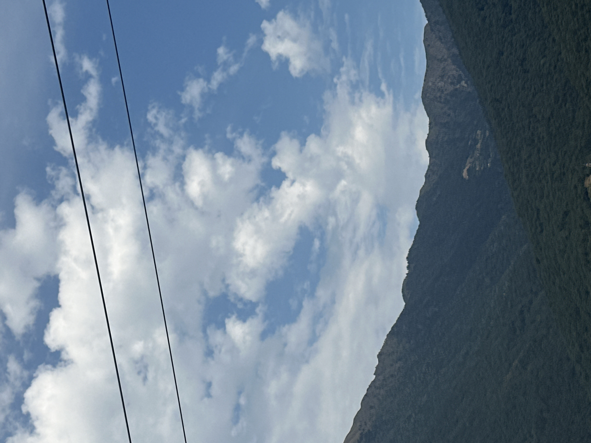 The image shows a view of mountains and sky, with two power lines crossing the frame horizontally in the upper section. The sky is blue with scattered white clouds. The mountains are covered in dark green foliage and appear rugged. The lower portion of the image shows a village nestled in the valley, with a church spire visible. The scene is likely a scenic view from the city of Artogne, Italy, given the mountainous terrain and architectural style.