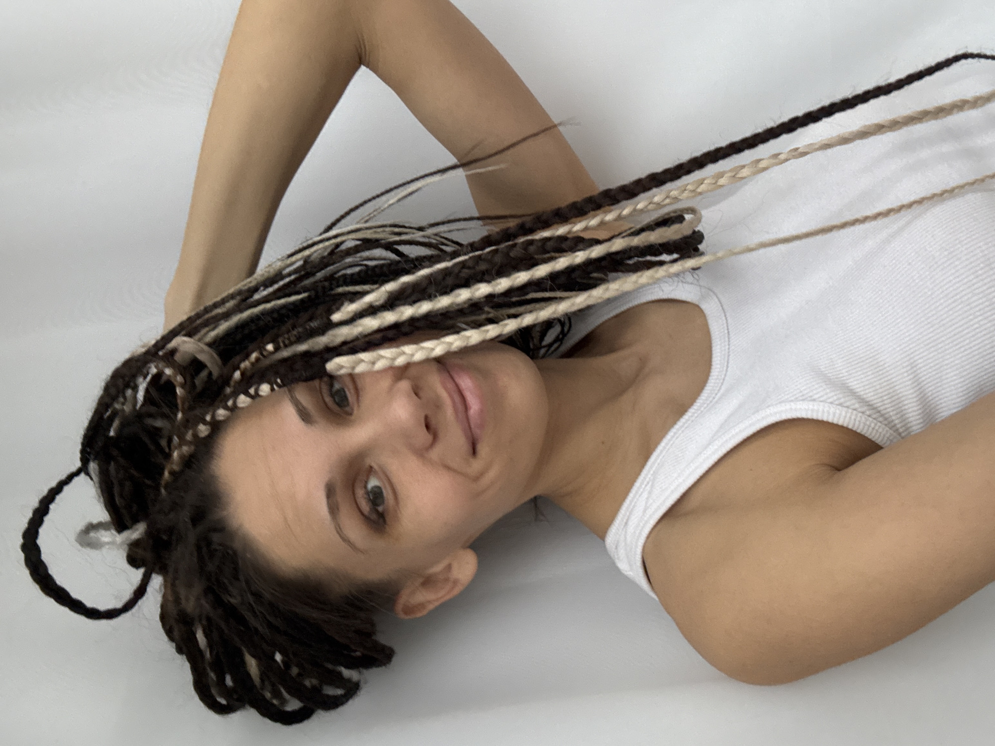 A woman is lying on a white surface. She has dark brown hair styled in braids with some blond braids intermixed. She is wearing a white tank top. Her right arm is bent and positioned above her head. The background appears to be a plain white surface. There is no information about the city of Rusanovo.