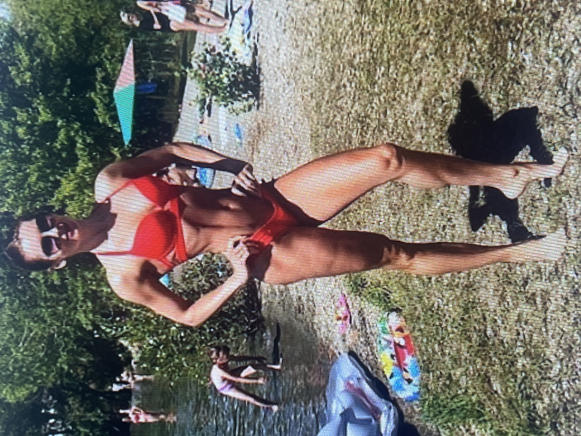 A woman in a red bikini poses on a grassy bank next to a body of water. She is wearing sunglasses and has her hands on her hips. In the background, other people are visible in the water and on the bank. There is a beach umbrella and some towels on the ground. The environment appears to be a park or recreational area on a sunny day. The location is not identifiable as Đà Nẵng, Vietnam.
