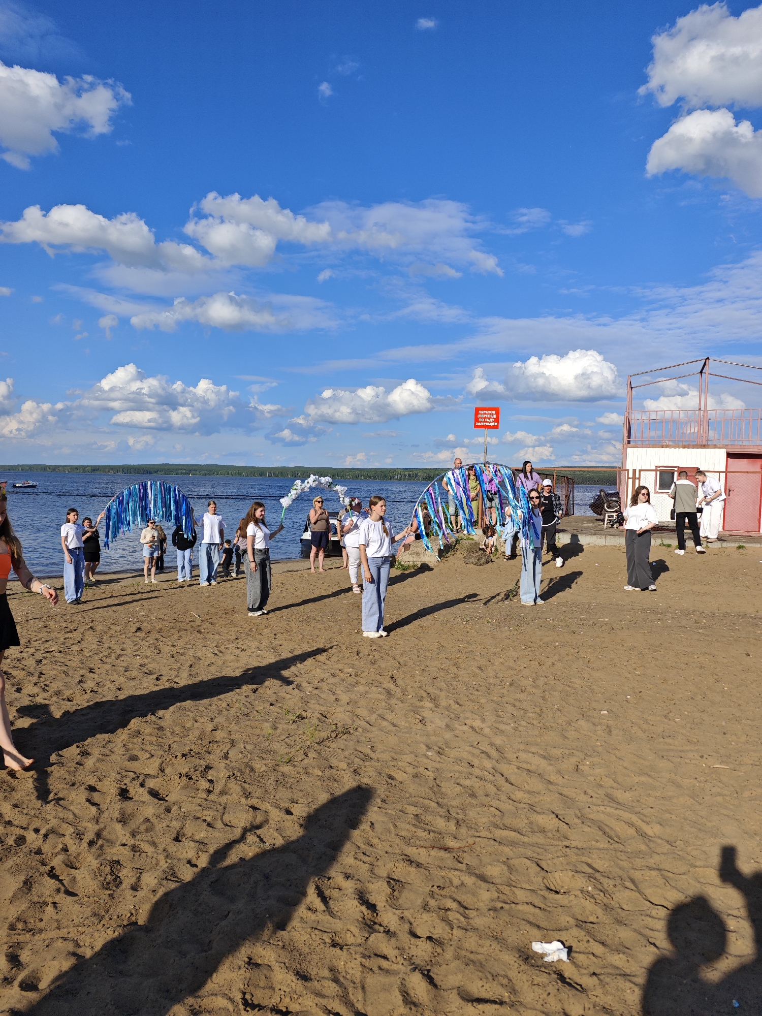 A group of young people are gathered on a sandy beach near the Kama River in Chaikovsky, Russia. A blue, arched structure decorated with blue and silver streamers is held up by several individuals, suggesting a festive or celebratory event. Other people in the group are holding white floral arrangements. In the background, a body of water stretches to the horizon, with a few boats visible. A building with a red roof and railing is located on the right side of the image, and a red sign with white lettering is posted on a pole. The sky is blue with scattered white clouds.