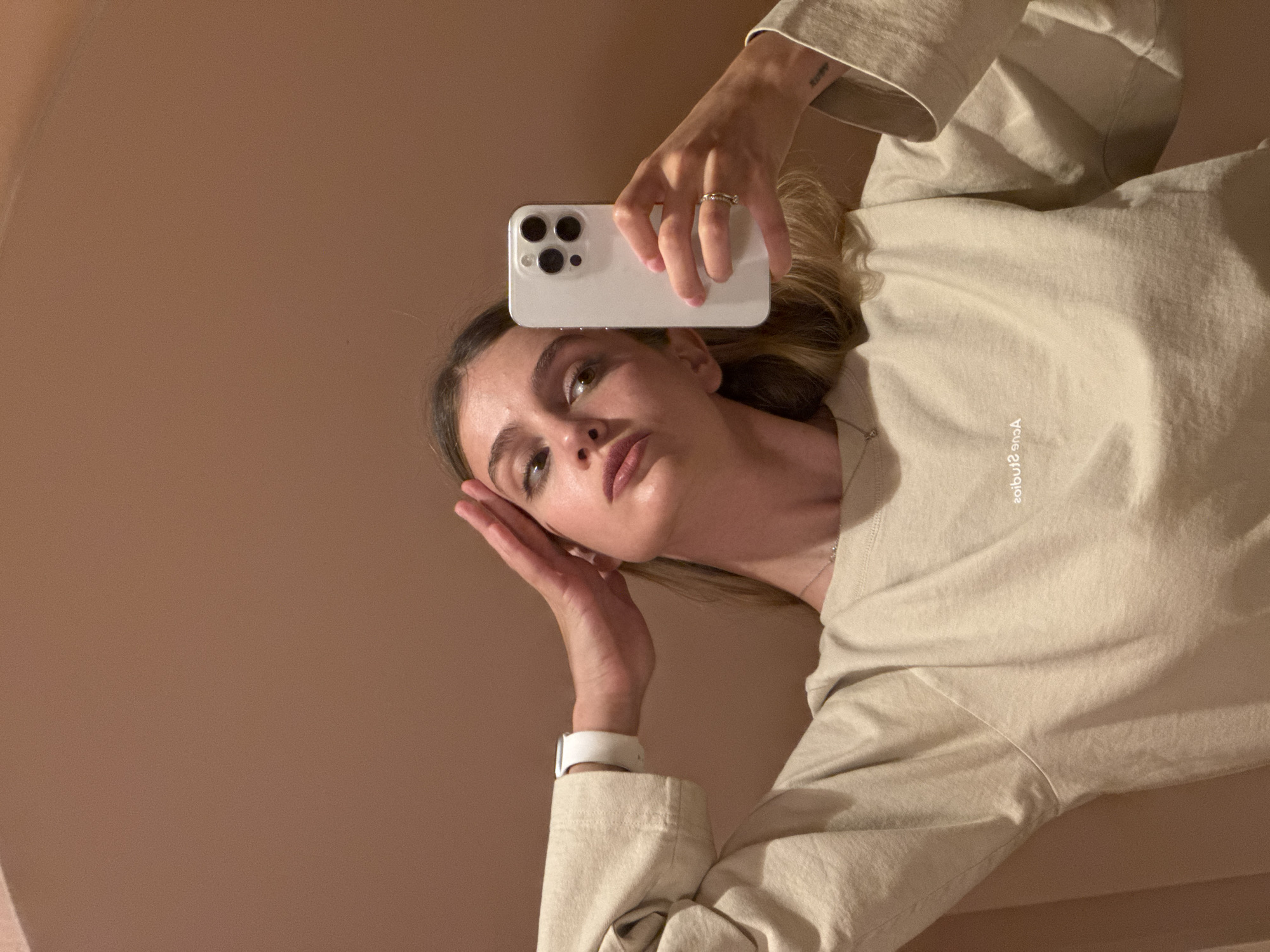 A young woman is taking a selfie with a white smartphone in a room with peach-colored walls. She is wearing a beige sweatshirt and a white smartwatch. Her hair is blonde. Her hand with a ring is holding the phone above her head. The scene's location in Tbilisi, Georgia, is not evident from the provided visual information.