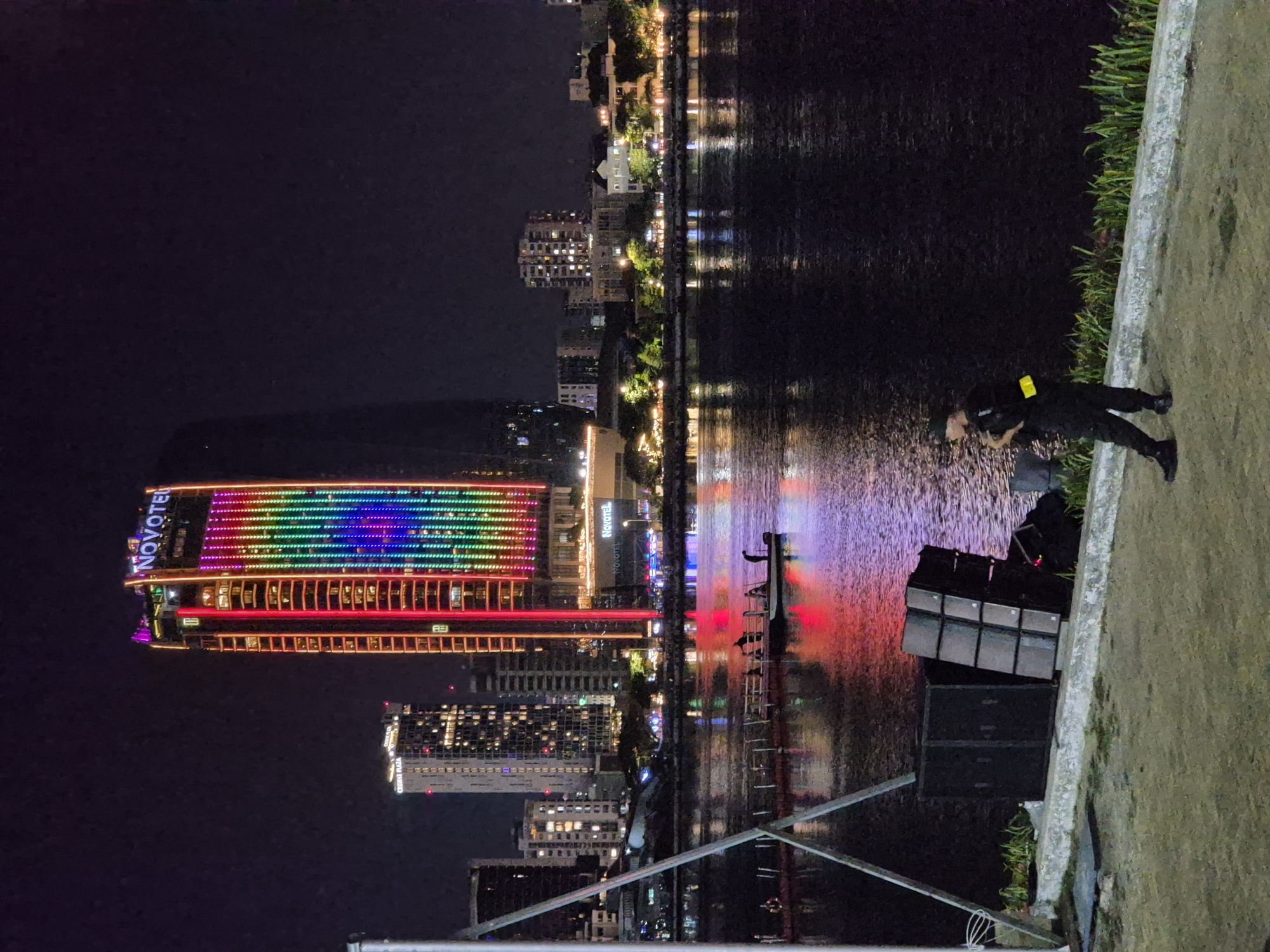 The image depicts a nighttime scene along the Han River in Da Nang, Vietnam. A prominent skyscraper, the Novotel Da Nang Premier Han River, dominates the skyline, its facade illuminated with a vibrant rainbow-colored LED display. Other buildings with illuminated windows are visible in the background, reflecting on the dark water. In the foreground, a man in dark clothing with a yellow armband stands near a stack of large black speaker cabinets. A small boat with several figures on board is moored on the river, with reflections of colorful lights on the water's surface. A metal structure, possibly part of a stage or scaffolding, is visible on the left side of the image. The overall atmosphere is urban and lively, characterized by the city's night lights and the presence of recreational activities on the river.