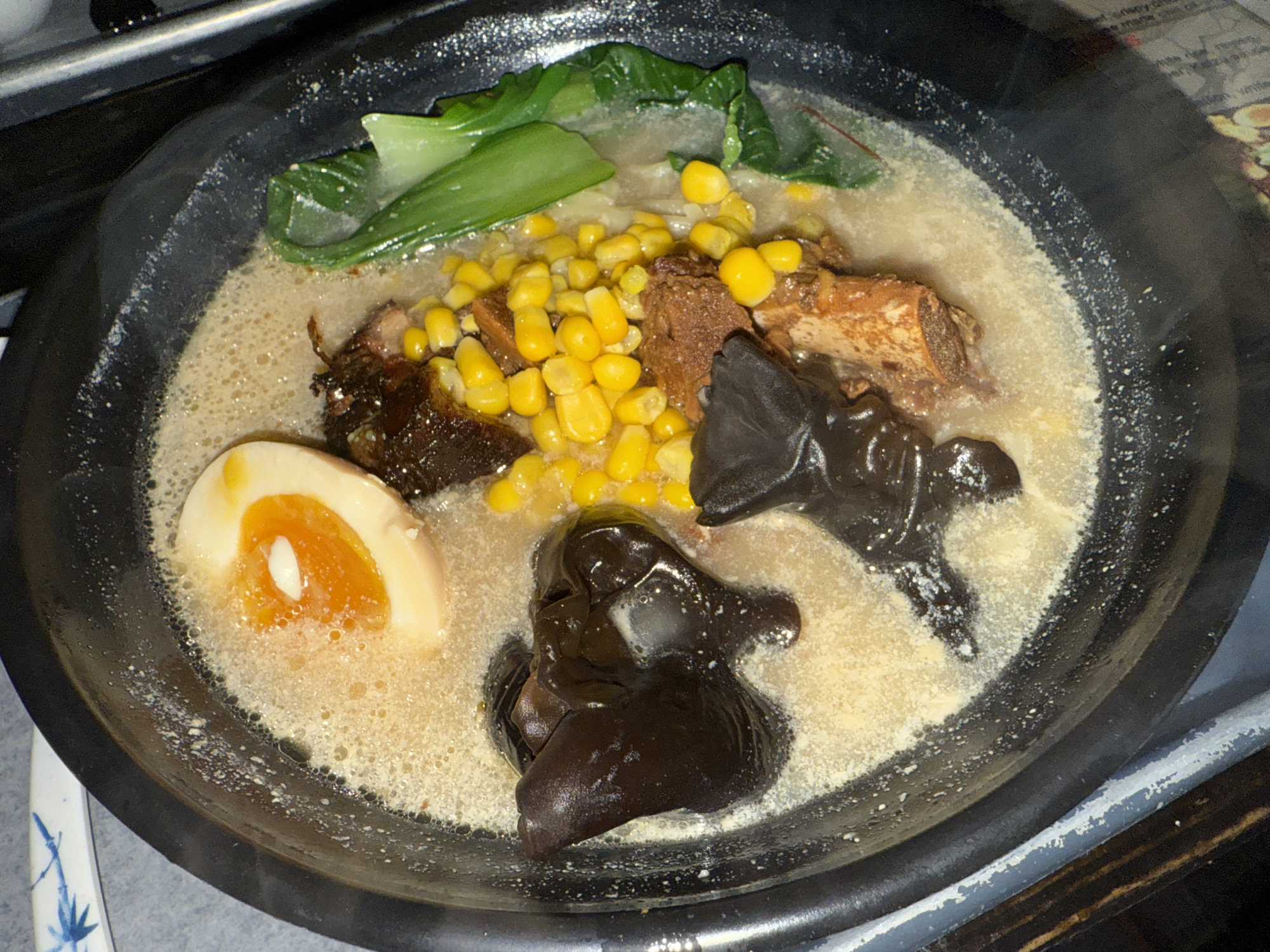 The image shows a bowl of ramen. The ingredients include: half a boiled egg, corn kernels, dark brown wood ear mushrooms, bok choy, ribs and broth. It is unknown whether the location is Dania Beach, United States of America.