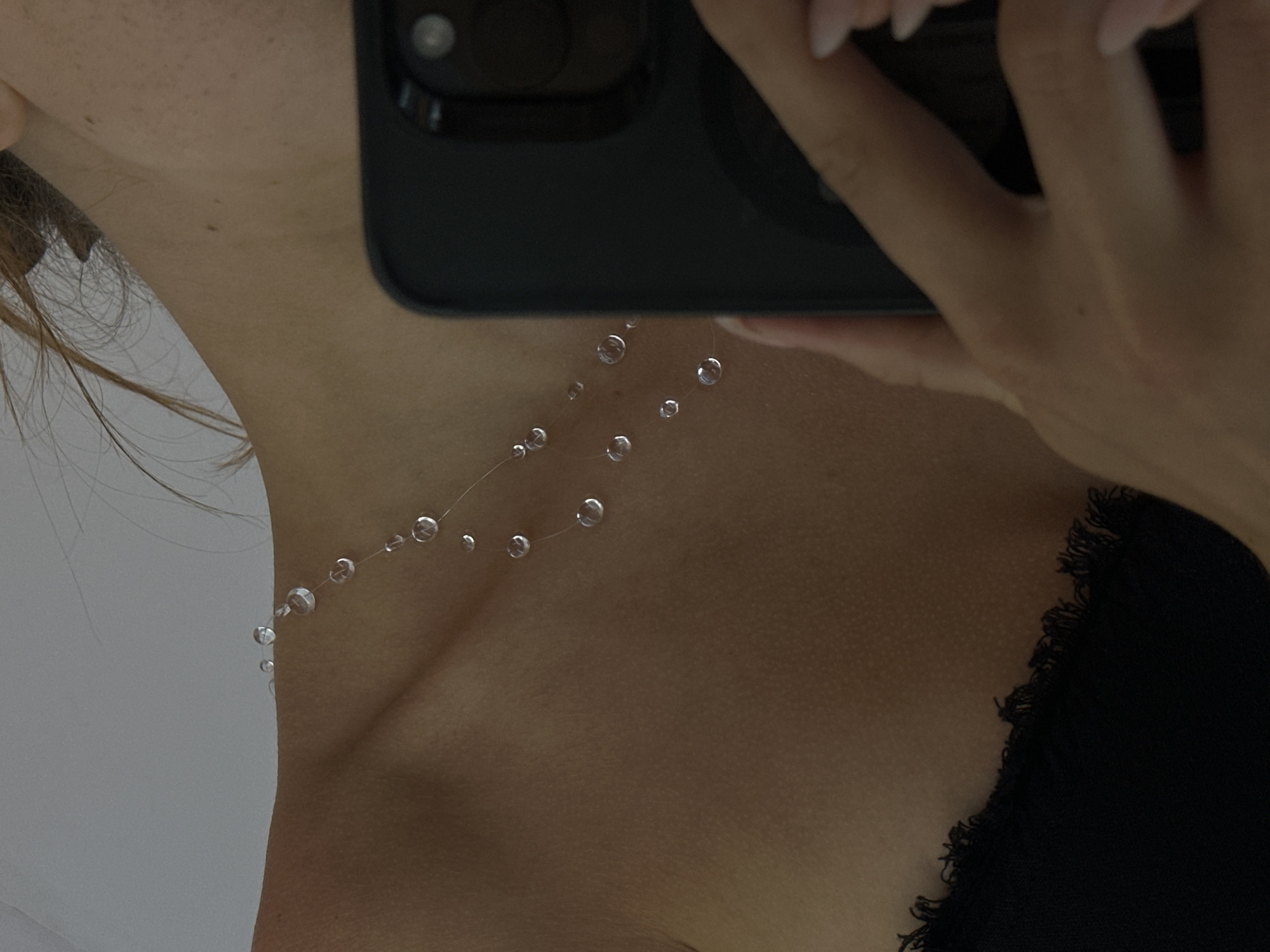 A close-up shot focuses on a person's neck and shoulder, where a delicate necklace with small, transparent beads is visible. The person is holding a black smartphone in front of their face, partially obscuring the view. The background is a plain white wall. The scene appears to be indoors, with soft lighting. No information suggests a specific location in North Kuta, Indonesia.