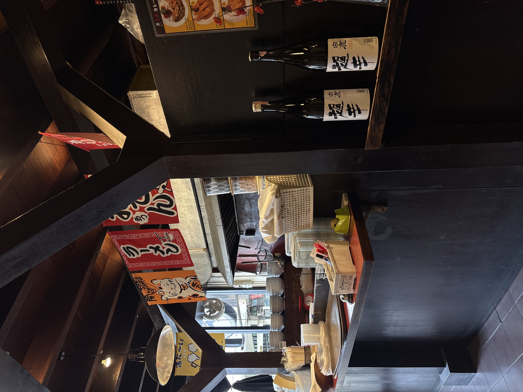 The image shows the interior of a Japanese restaurant, likely in Chiang Mai, Thailand. The scene is dimly lit, suggesting it might be evening or indoors with low lighting. The perspective is from below, looking up at wooden beams and shelves.
On a dark shelf, two bottles of sake are prominently displayed, labeled in Japanese script. Below the shelf, a variety of Japanese banners are hung, some featuring ramen and a lucky cat motif, adding to the cultural context. Other items visible include plastic containers, disposable cups, and what appears to be cutlery or chopsticks. In the background, there's a glimpse of a window showing the outside, and possibly other parts of the restaurant. The overall impression is of a cozy, traditional Japanese eatery with a focus on its offerings, suggested by the food images on a sign at the top. The text on the banners and bottles indicates Japanese cuisine and beverages.