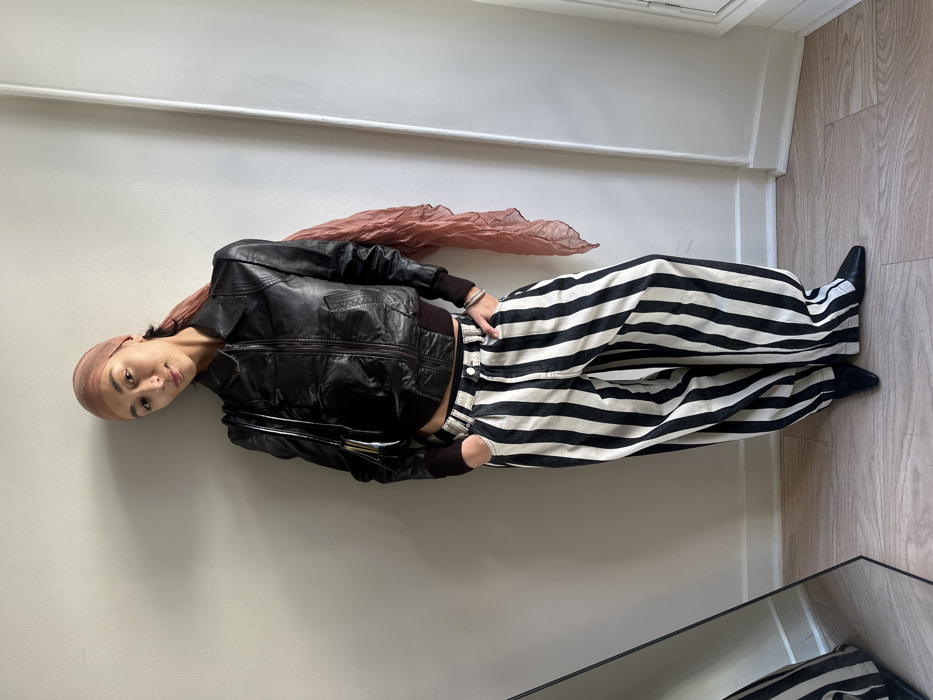 A female subject stands in a full-body pose within an interior room in Paris, France. The room features a plain, light-colored wall, a white baseboard, and light-toned wooden floorboards. A partial view of a floor-standing mirror is visible on the left, reflecting a portion of the subject's lower attire. On the right, a white vertical pipe or conduit extends along the wall. The subject wears a sheer, light reddish-brown headscarf over her dark hair, a black leather bomber jacket with ribbed cuffs and hem, and high-waisted, wide-leg trousers with vertical black and off-white stripes. A black shoulder bag with metallic accents hangs from her left shoulder, and a silver-toned bracelet is present on her left wrist. Her hands are placed into the front pockets of her trousers. She wears black pointed-toe flat shoes. She faces forward, her head slightly turned to the left, exhibiting a subtle smile.