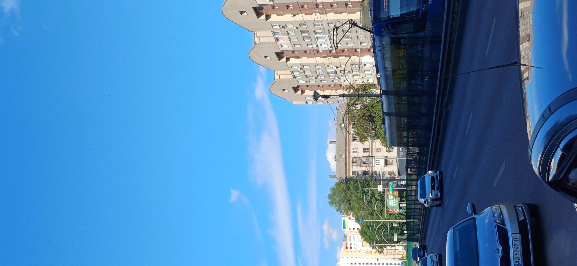 The media file shows a street scene in Kyiv, Ukraine, likely on a clear day. The image is oriented vertically, with the sky occupying the left side and the city street on the right.
The most prominent subjects are buildings and a tram. A tall, modern apartment building with a wavy roofline dominates the upper left portion of the frame, with many windows visible. To its right, a blue tram is moving along a track. Further down the street, older buildings with visible architectural details and windows are present, interspersed with trees and greenery. Several cars are driving on the road, including a white car in the foreground with a license plate that reads "AA 5747 TP". The sky is a vibrant blue with some wispy white clouds.
The time of day appears to be daytime, given the bright sky and shadows. The weather seems pleasant. The presence of the tram and the cars indicates daily urban activity. The Ukrainian flag is visible on one of the buildings in the background, reinforcing the Kyiv location. There is no discernible emotion conveyed in the scene.