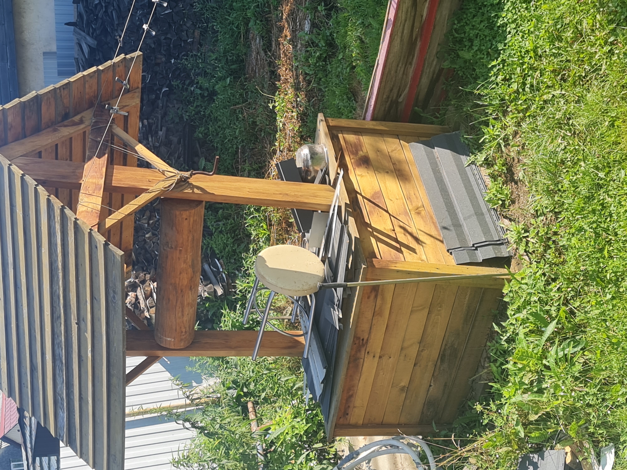 This image captures a rustic wooden well structure situated outdoors, likely in a residential area or a dacha (summer house) property in Angarsk, Russia. The well has a pitched roof made of wooden planks, supported by sturdy wooden posts. A large, weathered log is integrated into the structure, serving as a pulley mechanism. The base of the well is a wooden box, also constructed from planks.
On top of the well's base, various items are piled, including what appears to be roofing material, a metal frame, and a round, beige object that might be a lampshade or a weather vane component. A glass jar with some contents is also visible. The surrounding environment is overgrown with lush green grass and various weeds, suggesting a semi-wild or unkempt garden setting. In the background, a stack of firewood is visible, and hints of buildings with tiled roofs can be seen. The overall impression is of a functional, albeit somewhat cluttered, outdoor space in a rural or suburban Russian setting.