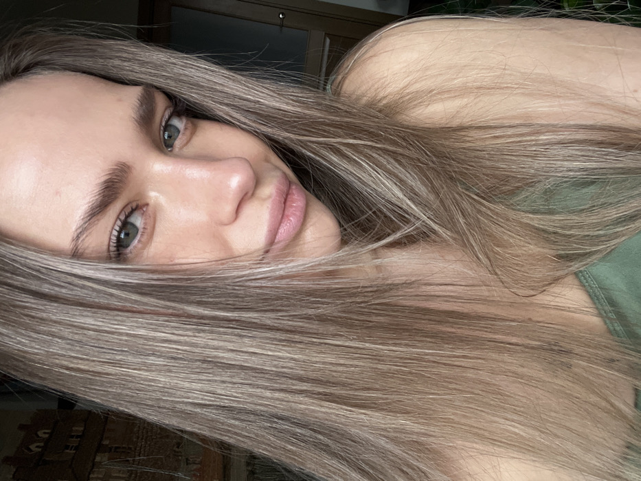 A close-up portrait depicts a young adult female with light skin and long, straight, light brown to blonde hair, centrally parted and framing her face. Her eyes are green-blue, and her naturally full lips exhibit a faint pink hue, curved into a subtle smile or smirk. She wears an olive-green tank top or camisole, visible on her upper chest and right shoulder. The subject maintains direct eye contact with the viewer. The background indicates an indoor environment. Visible elements include a partial view of a wall hanging or framed artwork with architectural motifs in muted colors on the upper left, and a light-colored wooden door and frame on the right. These interior features are generic and do not provide specific visual cues for the forensic identification of a precise location within Saint Petersburg, Russia.