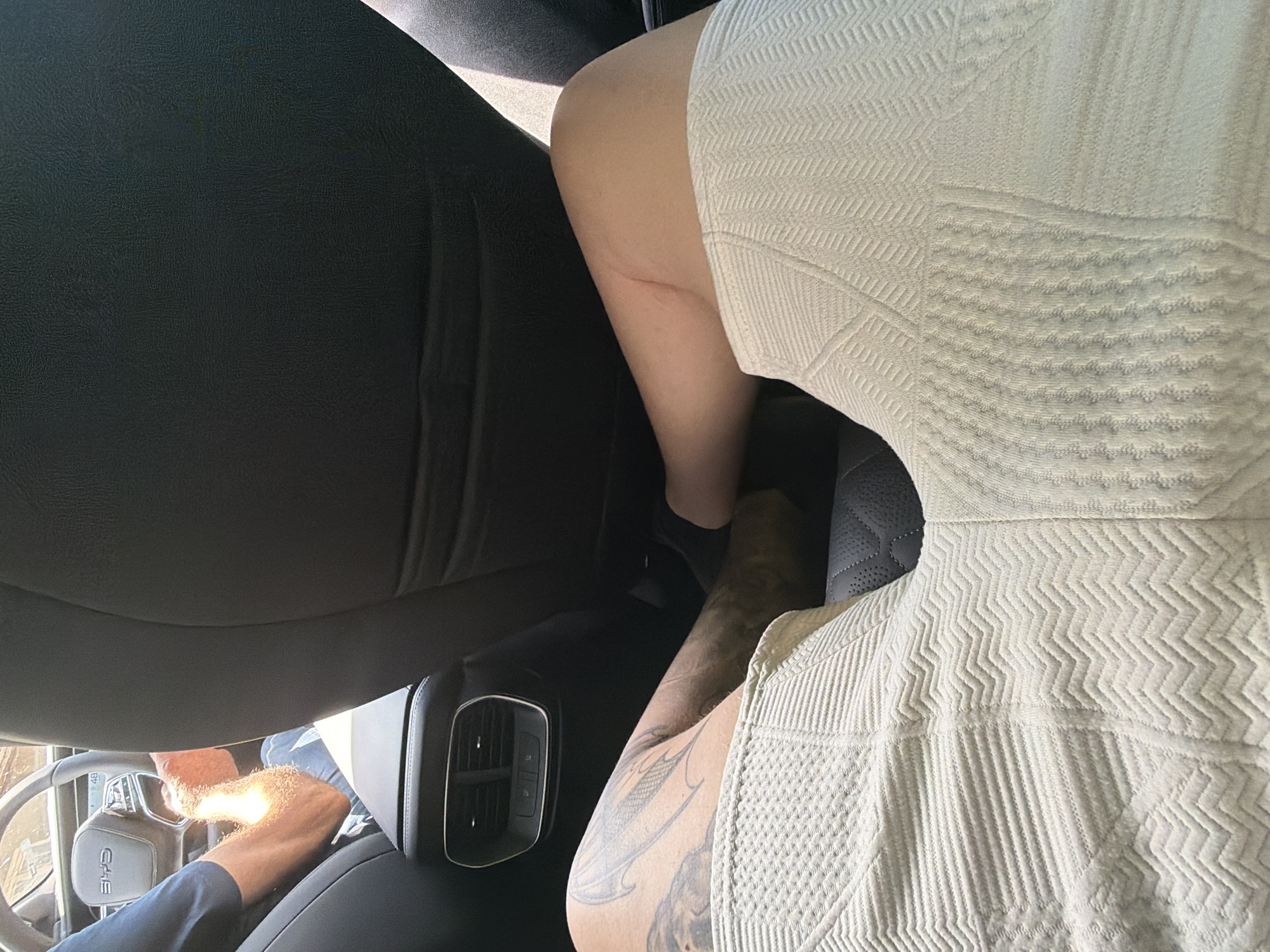 The image captures a close-up view of the interior of a car, taken from the backseat. In the foreground, a person's legs are visible, clad in a light-colored, textured garment. Their legs are positioned across the frame, with tattoos visible on one leg. To the left, the steering wheel and dashboard of the car are partially visible, suggesting a driver is present. Sunlight streams into the car, illuminating the driver's arm. In the background, the back of the car seats and air vents are discernible. The overall scene suggests a journey within a vehicle, likely in a car, with no specific indicators of the city of Dolno Ablanovo, Bulgaria.