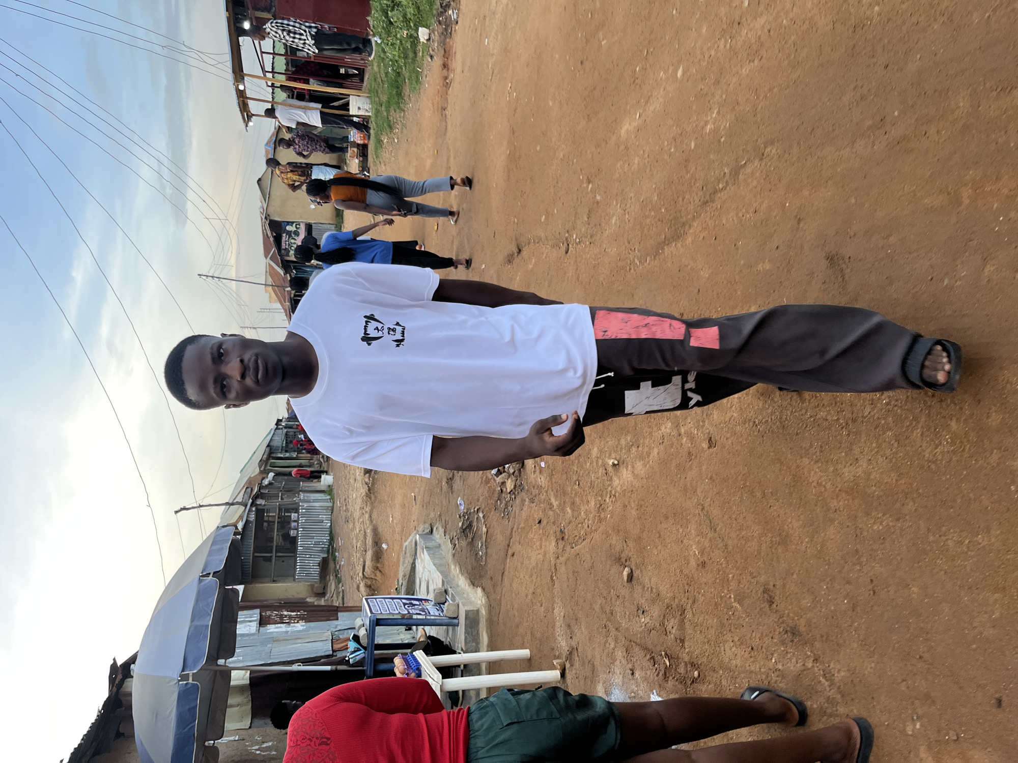 The image presents a lively street scene in Jos, Nigeria, featuring a young man as the primary subject. He stands in the foreground, directly facing the camera, with short dark hair and a serious expression. He wears a white t-shirt with a black graphic on the chest, which appears to be a stylized animal head (possibly a lion or bear) with some indistinct text beneath it, and dark grey track pants with a prominent red stripe running down the side. He completes his attire with black flip-flops.
The setting is an unpaved, reddish-brown dirt road, characteristic of many local markets or residential streets in the region. The time of day appears to be daytime, possibly late afternoon or early evening, given the soft, slightly overcast sky. Numerous power lines stretch across the upper left portion of the sky.
In the background, the street is bustling with other pedestrians. A person in a red top and dark shorts is partially visible in the lower left, seated near a blue and grey striped market umbrella, with a blue table and an illegible white-on-blue sign beside them. Further back, individuals in a blue shirt and an orange top are seen walking, suggesting a flow of daily activity. The sides of the street are lined with rudimentary structures, likely market stalls or small shops, constructed from corrugated metal sheets and wood, contributing to the informal market atmosphere common in Jos.