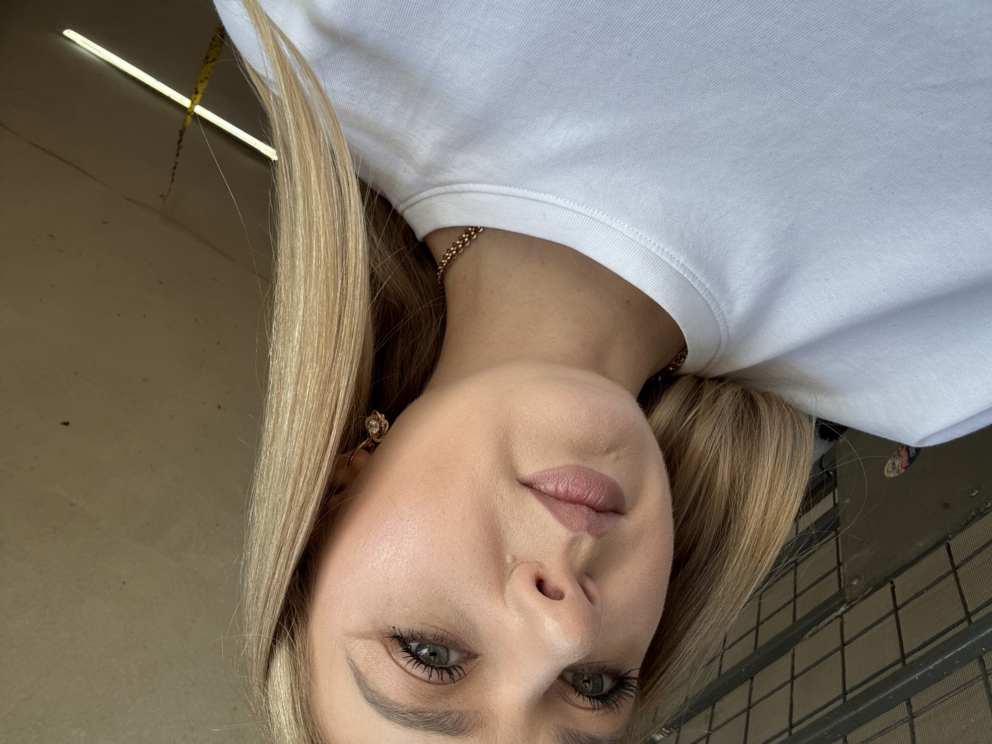 A young blonde woman, positioned in the lower right portion of the frame, is looking directly at the camera. She is wearing a white t-shirt and a gold chain necklace. The background consists of a plain, textured wall.