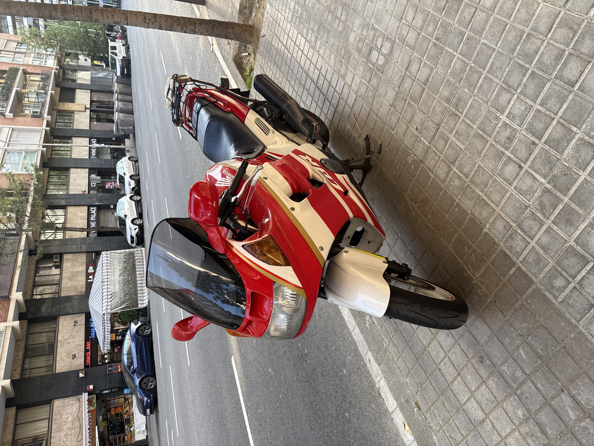 A red and white Honda CBR600F motorcycle is parked on a gray-tiled sidewalk next to a street in Barcelona, Spain. The motorcycle is facing the viewer. The bike has a black windscreen, a black seat, and a luggage rack. The building in the background features businesses with signs in English, including "WINE PALACE" and "SNACK BAR LA LUR". Several cars are parked along the street, and a tree is visible next to the bike.