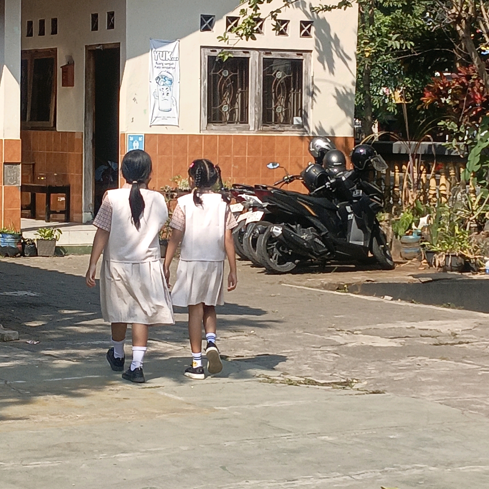 Two young girls in school uniforms walk away from the camera on a paved path in front of a building. The girl on the left has her dark hair in a ponytail, and the girl on the right has her dark hair in two braids with pink bows. Both girls wear white shirts with plaid collars and white sleeveless vests, and white skirts. They are also wearing white socks and dark shoes. Behind the girls are parked motorcycles with helmets on them. The building behind them has tiled walls, windows with decorative metal bars, and a banner hanging on the wall with the text "YUK..." and a cartoon image of a sad cup. This scene is likely in Salatiga, Indonesia, as indicated by the context. The overall environment appears to be an outdoor school or community area on a sunny day.