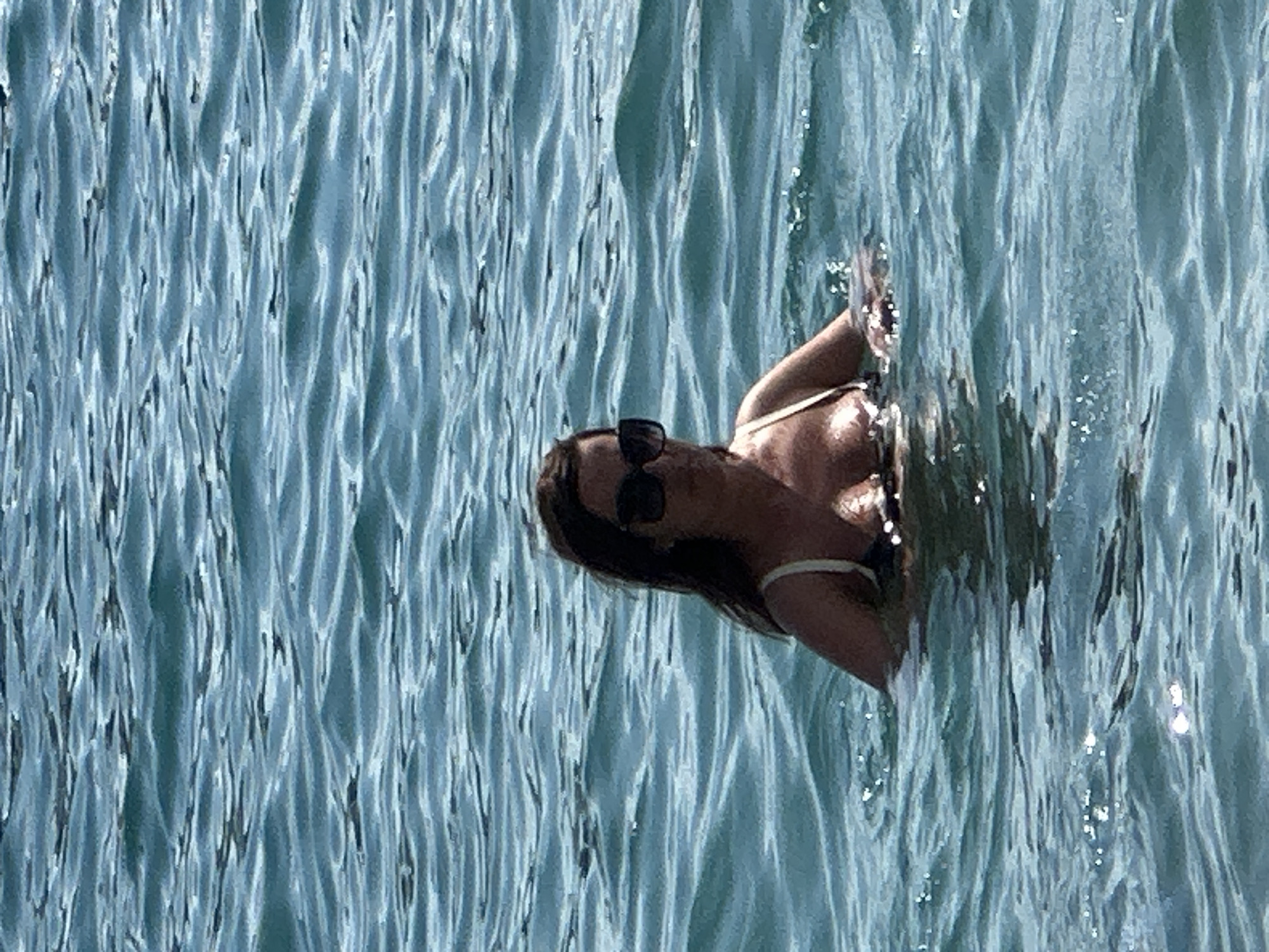A woman swims in the clear, blue water of the sea off the coast of Budva, Montenegro. The sun is shining, reflecting off the rippling surface of the water. The woman is wearing a bikini and sunglasses, and she is looking down at the water. The scene is peaceful and serene, with the sound of the waves lapping against the shore. The time of day is likely midday, given the bright sunlight. The water appears calm, suggesting good weather conditions. The woman's posture and gaze suggest a relaxed and contemplative mood. There is no visible text in the image.