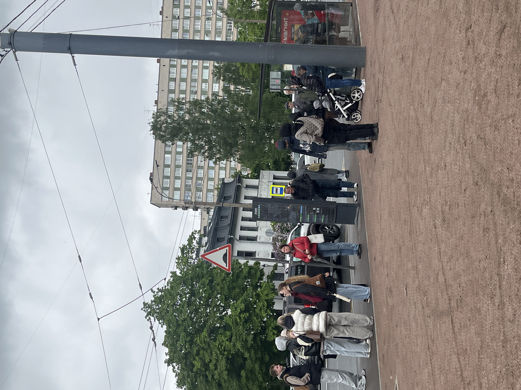 The image shows a street scene in Saint Petersburg, Russia. A group of people are waiting at a bus stop next to a pedestrian crossing with a "Give Way" road sign visible. The bus stop is adjacent to a sidewalk covered in light brown gravel. Overhead power lines and supporting poles are visible in the foreground. Behind the people waiting are buildings, trees, and parked vehicles. There is a bus shelter with advertisements behind the people waiting for the bus.