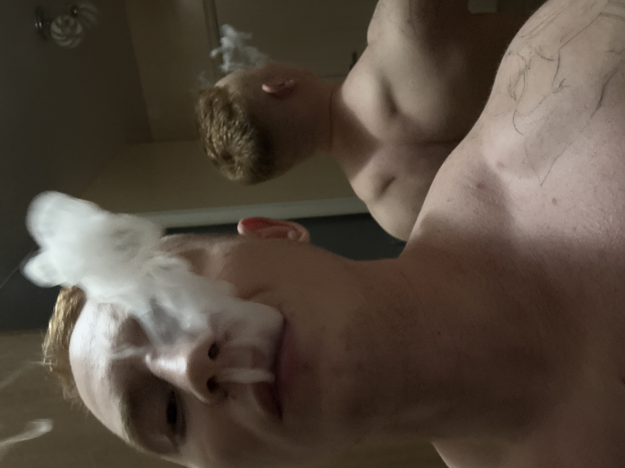 The image depicts two shirtless Caucasian males in an indoor setting. The foreground individual is facing the camera, exhaling a cloud of white smoke from their nose and mouth. Their hair is blonde. The person behind has auburn hair and also exhales smoke. The ceiling and a light fixture are visible, but no verifiable location data is available to conclusively place the image in Chaikovsky, Russia.