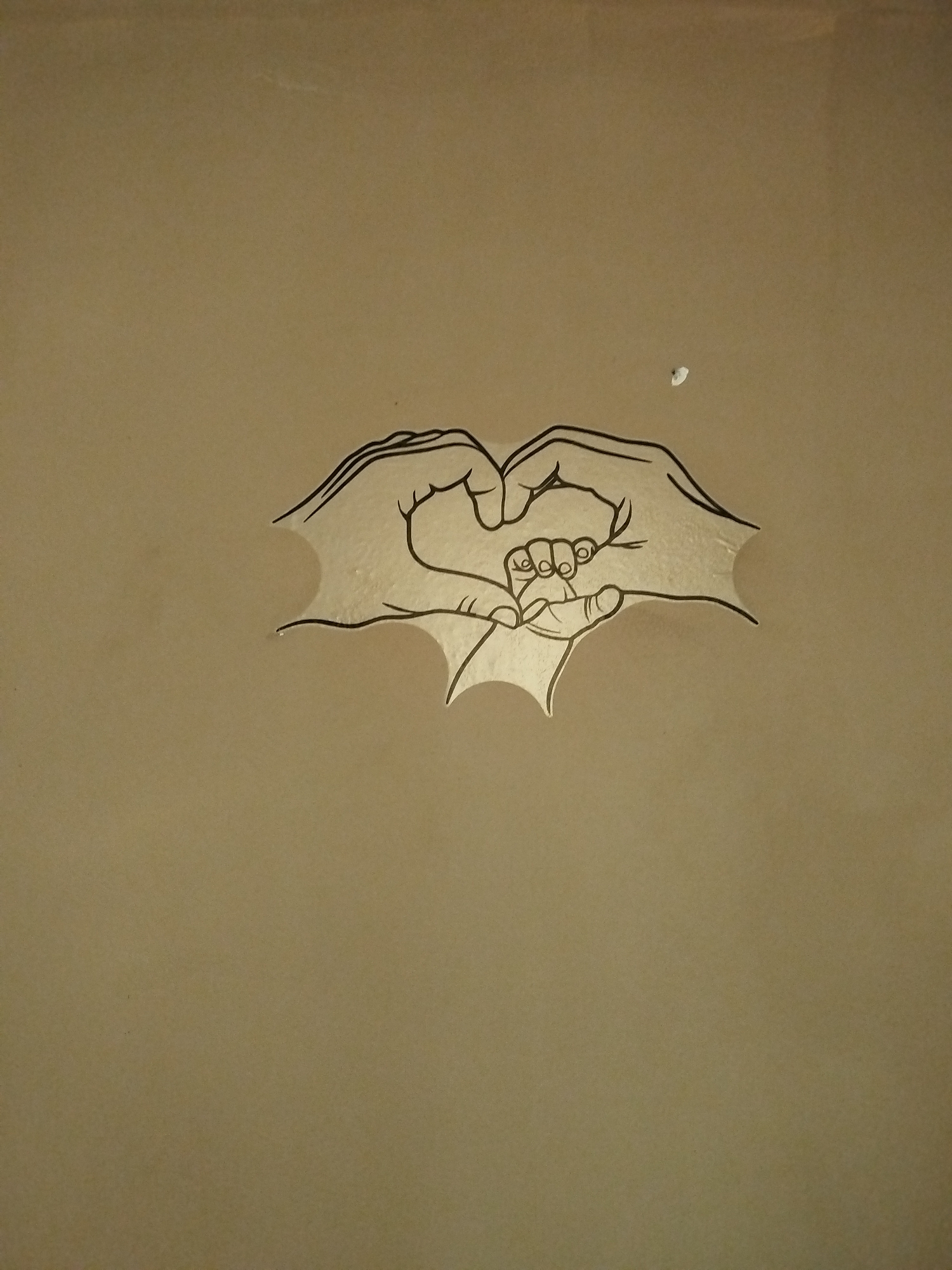 The image shows a drawing of two adult hands forming a heart shape around a smaller baby's hand. The drawing is on a tan or light brown surface, possibly a wall. A small white mark is visible near the top of the image. The location is unknown.