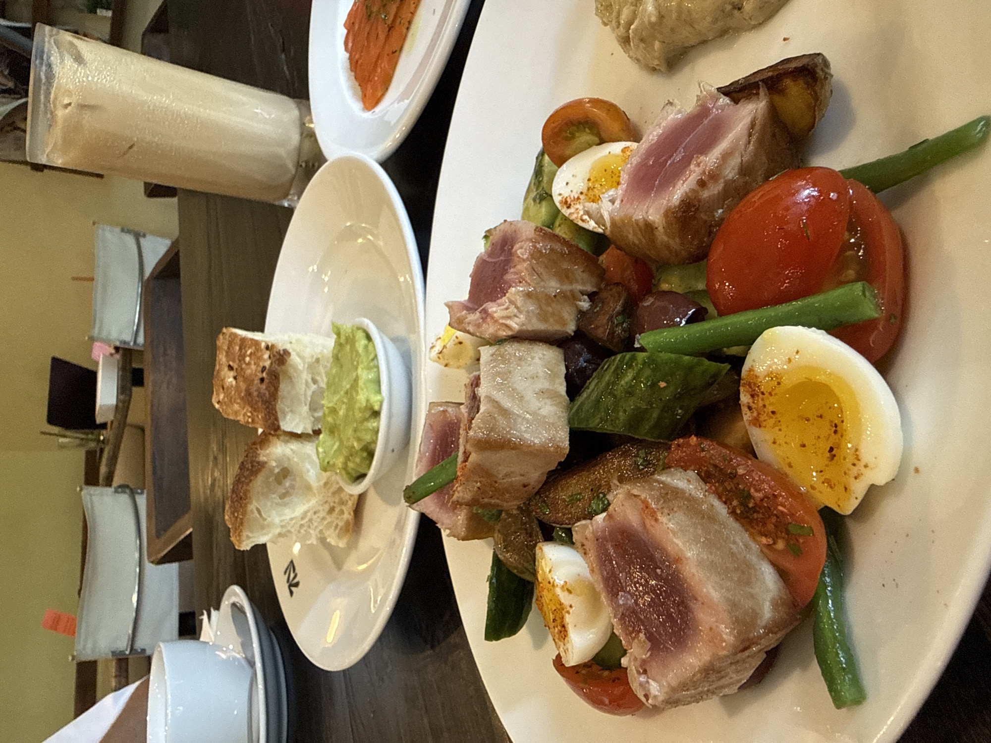 This image captures a vibrant culinary spread set on a dark wooden table, likely within a restaurant or cafe in North Kuta, Indonesia. The main focus is a large white plate presenting a colorful salad, reminiscent of a Nicoise salad. It features thick slices of seared tuna, still pink in the center, alongside halved bright red tomatoes, quartered hard-boiled eggs with a sprinkle of seasoning, green beans, and what appear to be dark olives and possibly roasted potatoes or bread cubes.
To the left, a smaller white plate holds several rustic slices of bread, possibly sourdough or seeded, served with a small bowl of a green, creamy spread, which could be guacamole or a pesto. Further left, partially visible, is another white plate with thin slices of orange-hued fish, likely smoked salmon.
In the upper left corner, a tall glass contains a light-colored, frothy drink, possibly a smoothie or milkshake. Below the bread plate, a white coffee cup and saucer are visible. In the background, white chairs with simple designs are partially seen, suggesting an indoor dining environment. The overall lighting is warm, enhancing the presentation of the food. On the small bread plate, a subtle, stylized logo appears, possibly resembling the letters "RK". No people are visible, but the arrangement implies a meal either being served or recently enjoyed.